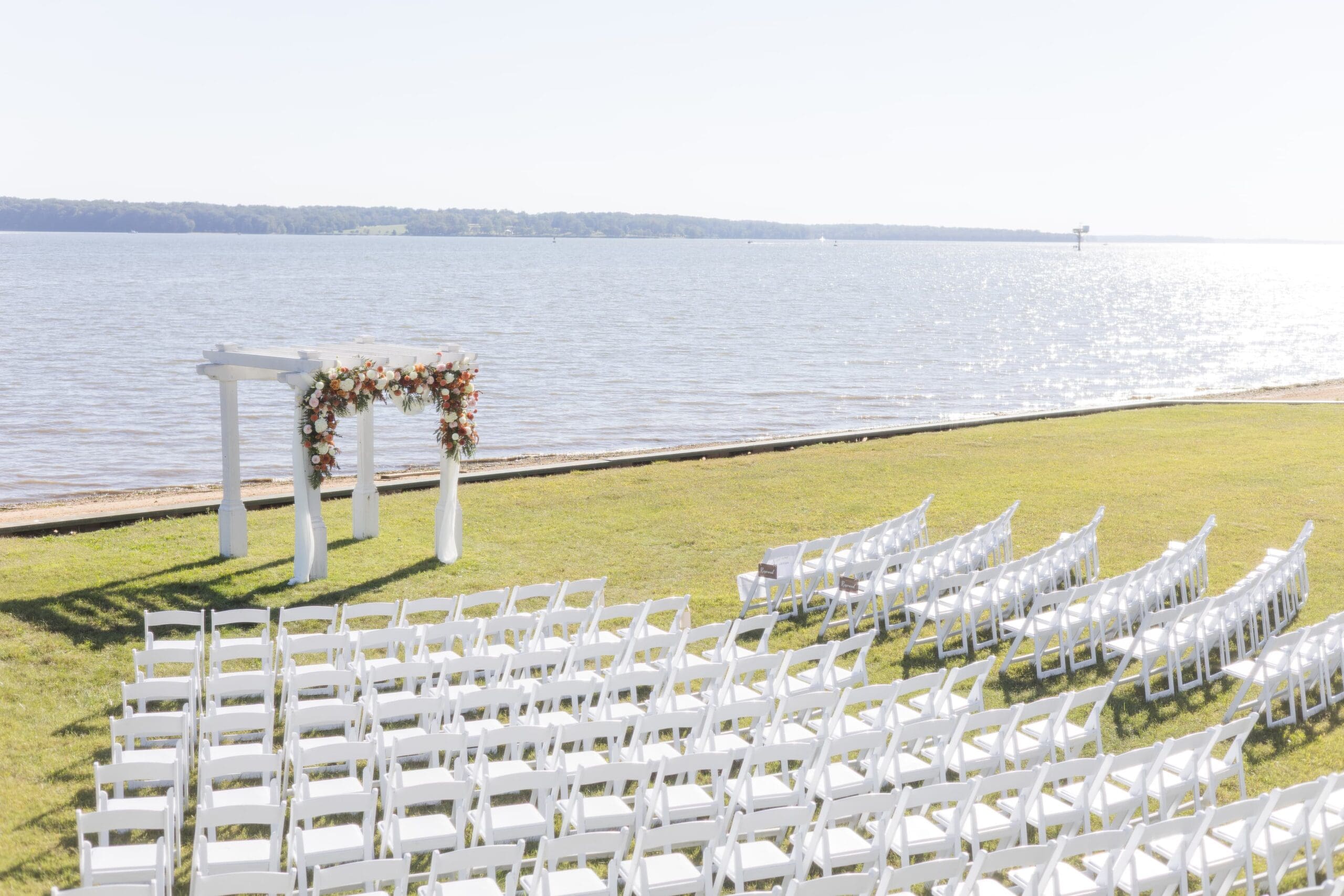 Outdoor wedding setup by a lake, featuring rows of white chairs on green grass, a floral arch, all under a clear blue sky.