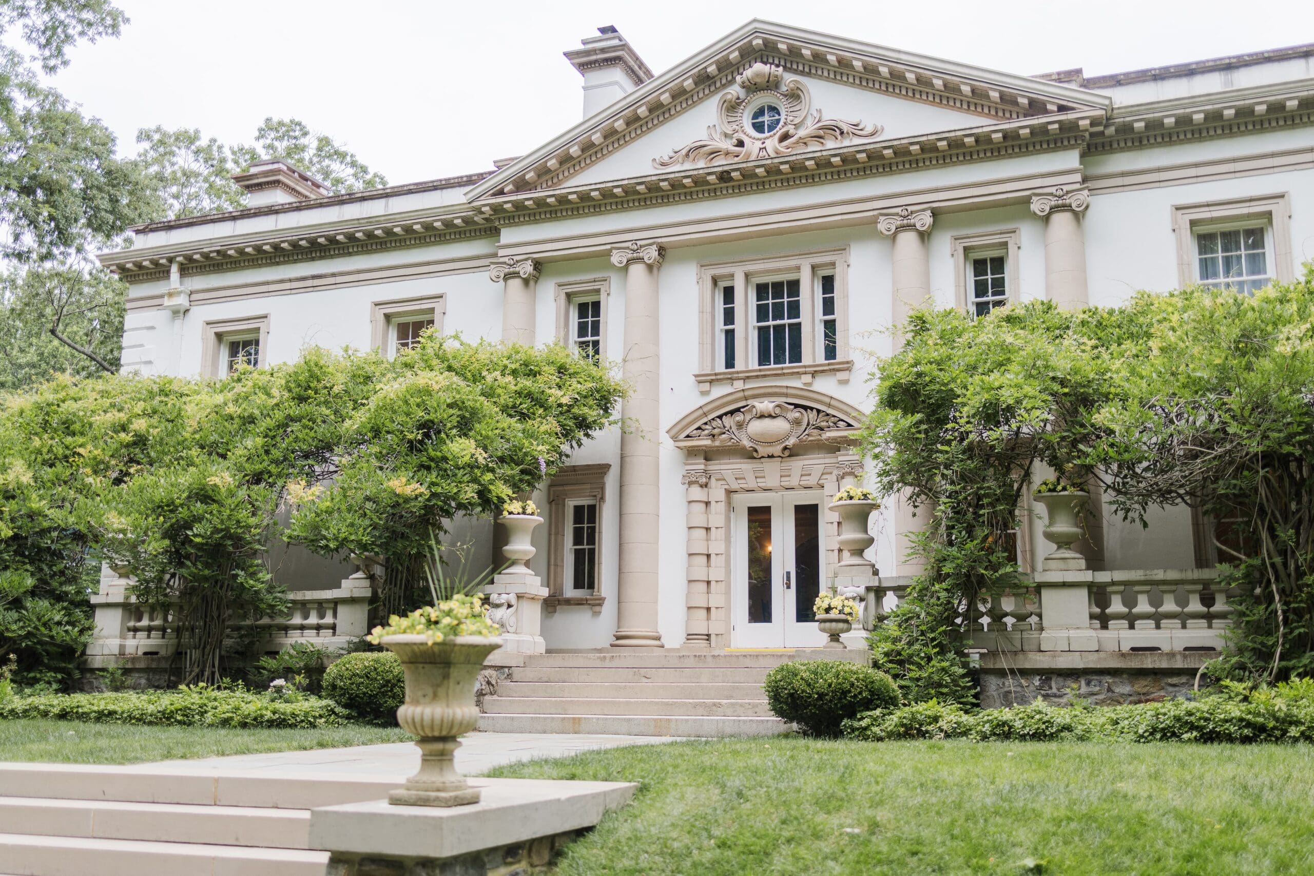 Elegant white mansion with wisteria-covered walls, large porch. Surrounded by lush greenery and manicured bushes.