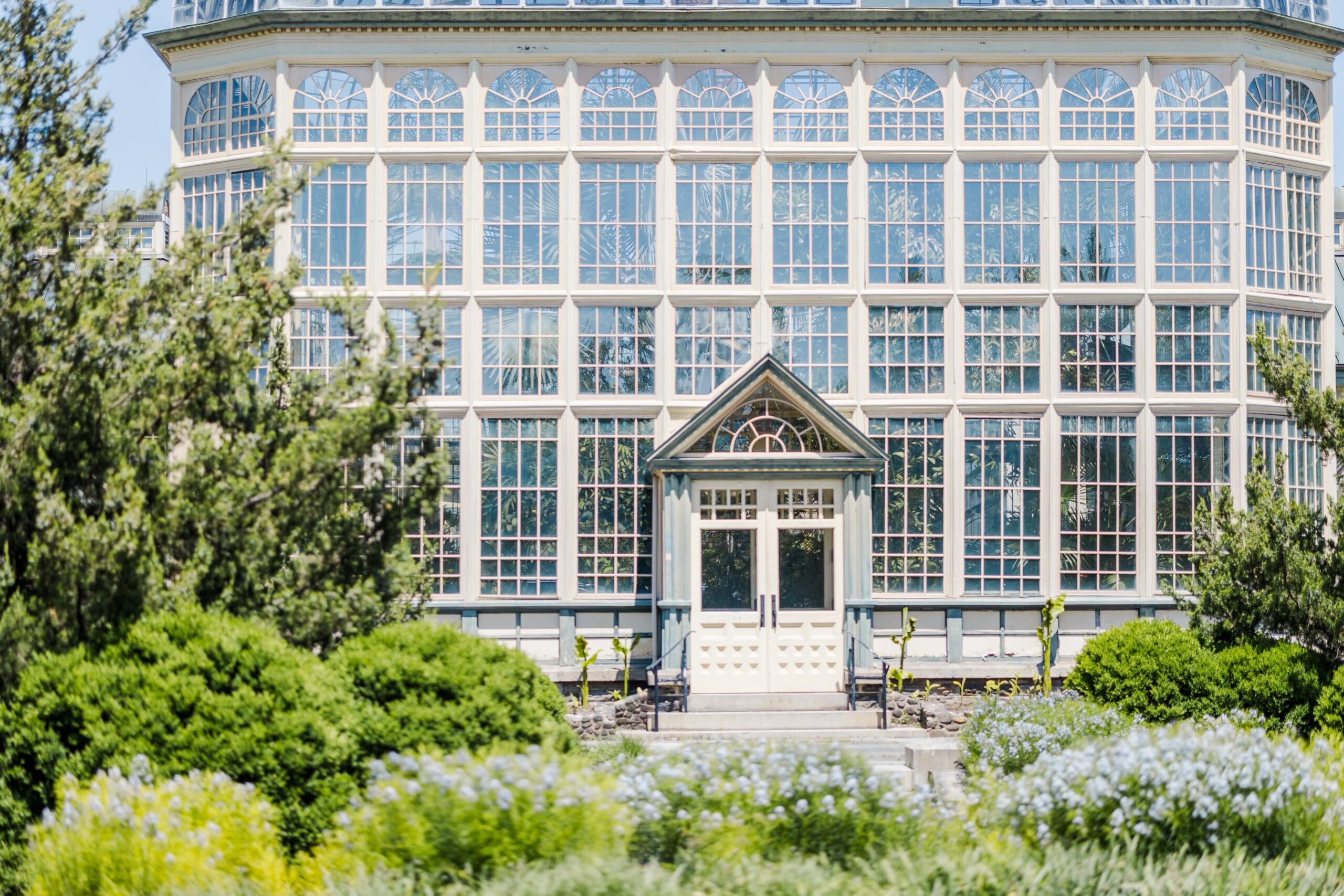 A large, glass-paneled greenhouse with a classic facade is surrounded by lush green shrubs and colorful flowers, under a clear, sunny sky.