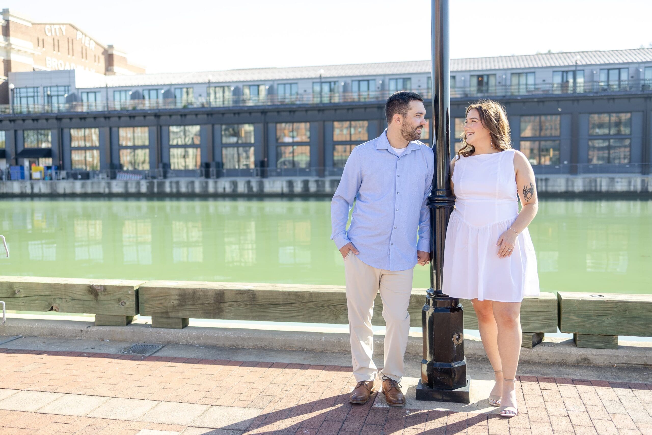woman in white dress leans against a light post as her fiancé leans against the other side smiling at each other