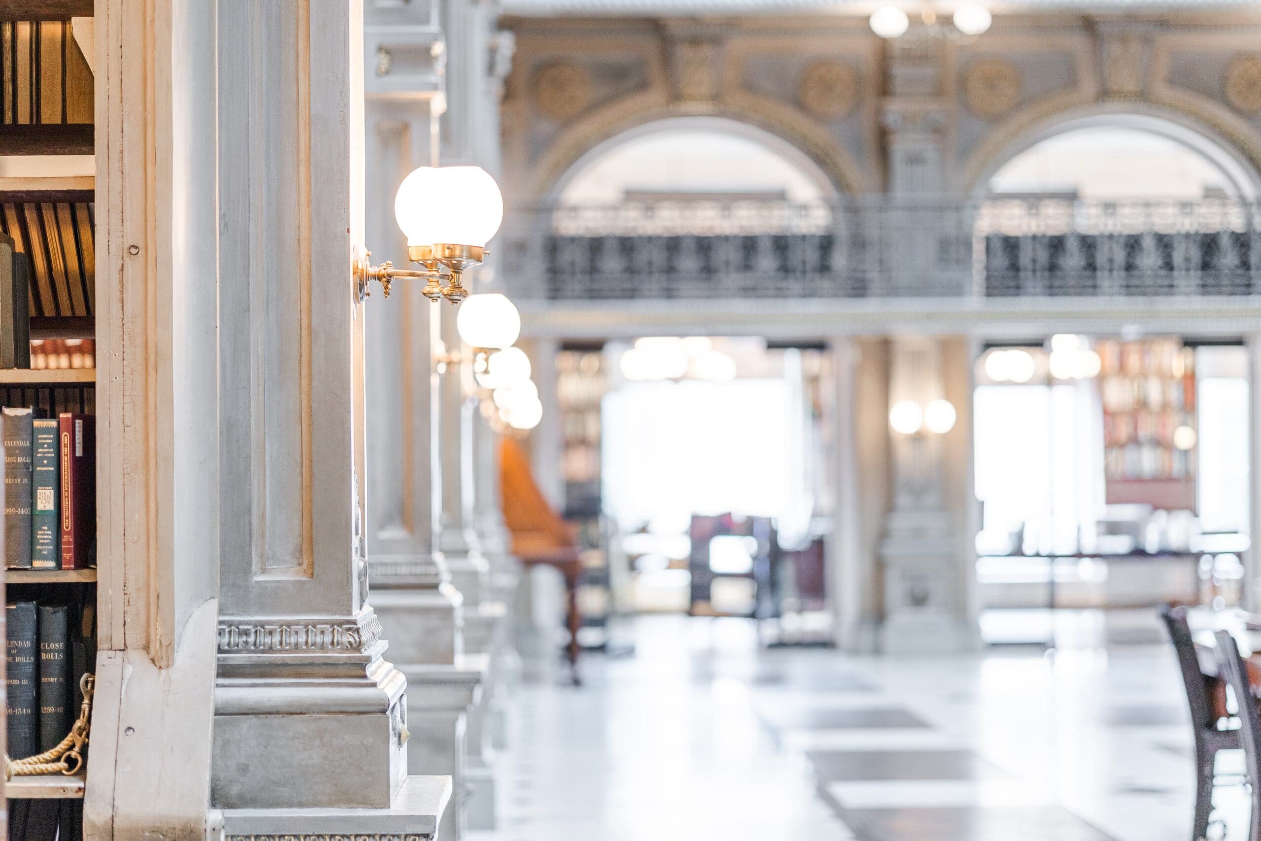 Bright, elegant library interior with tall columns, glowing wall lamps, and bookshelves lining the aisle. The atmosphere is serene and classic.