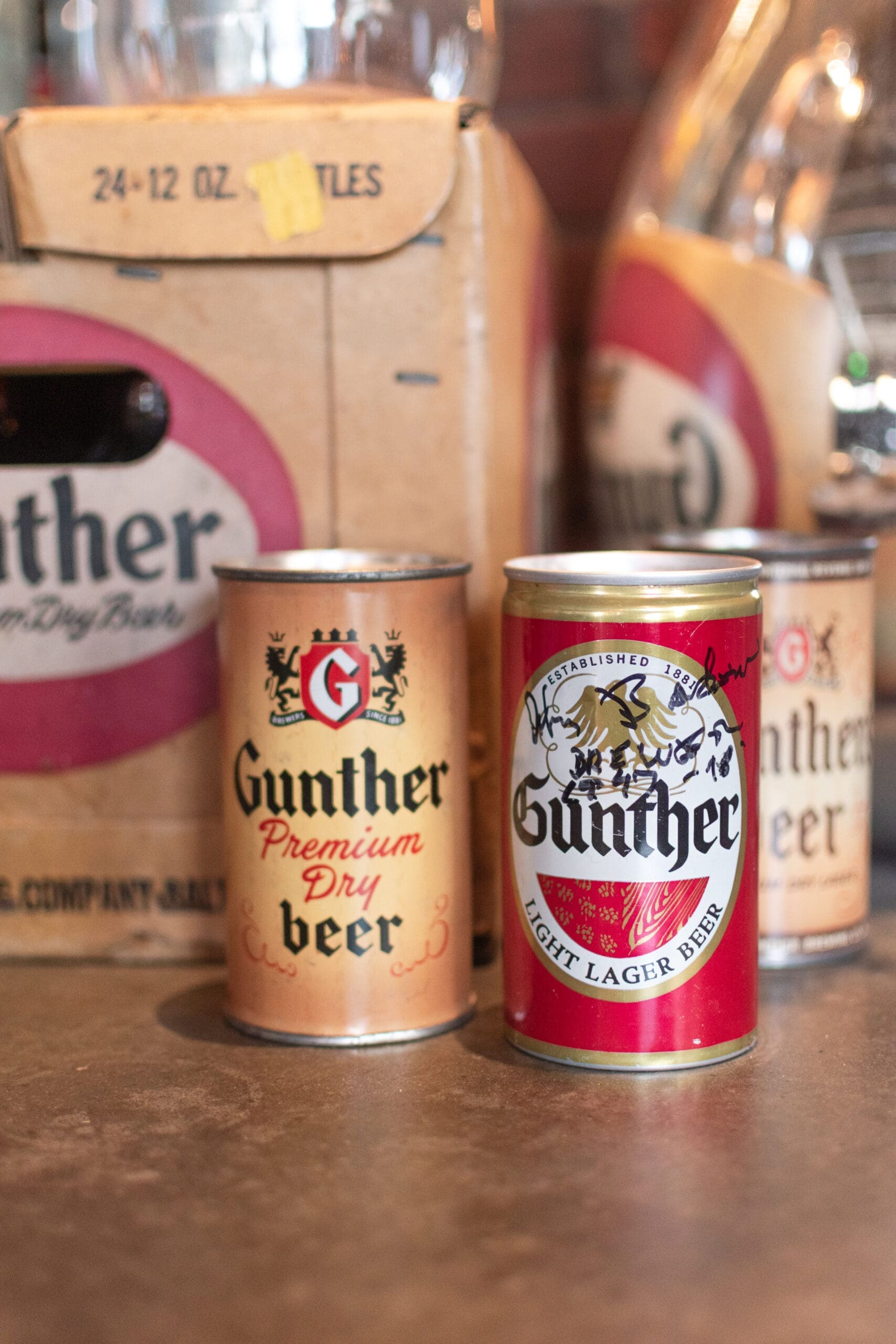 Cans of Gunther beer are displayed next to a cardboard box with a vintage logo. The scene conveys a nostalgic, retro vibe.