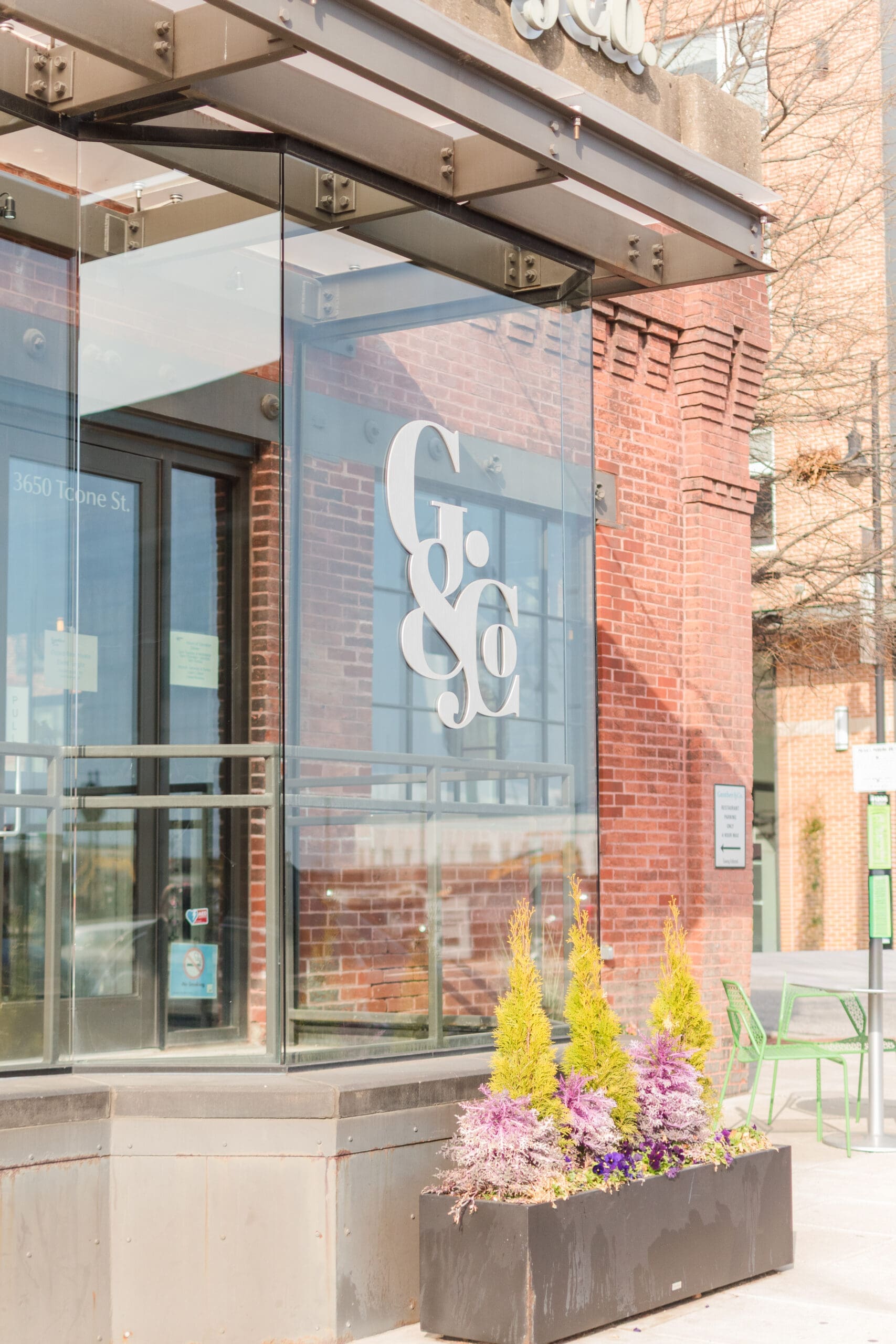 Glass entrance with large white "G&C" logo on a modern building with red bricks. Yellow flowers are in the foreground, adding a cheerful touch.