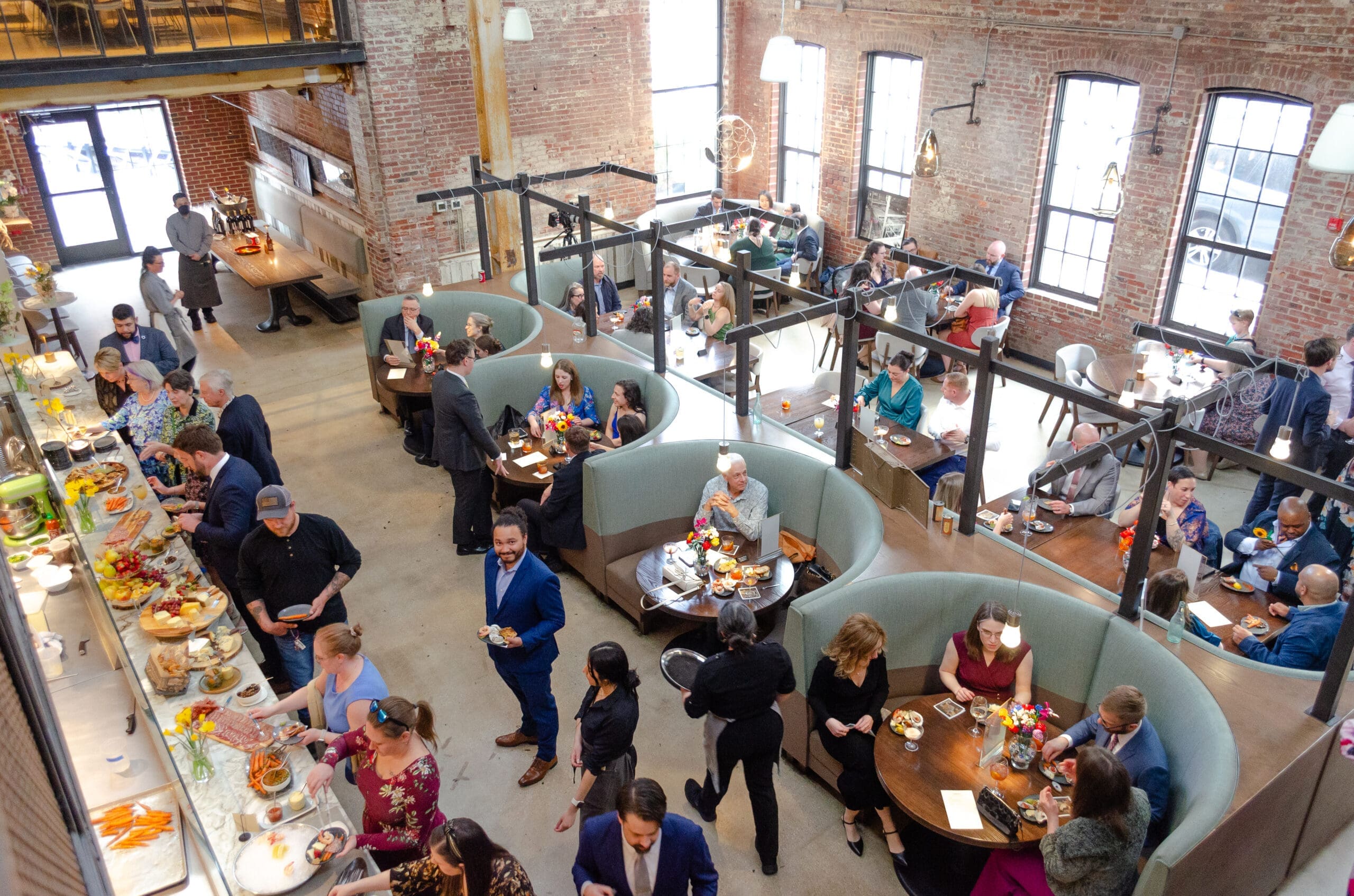 A bustling restaurant scene with diners seated in curved booths and tables. Exposed brick walls and large windows create an airy, lively atmosphere.