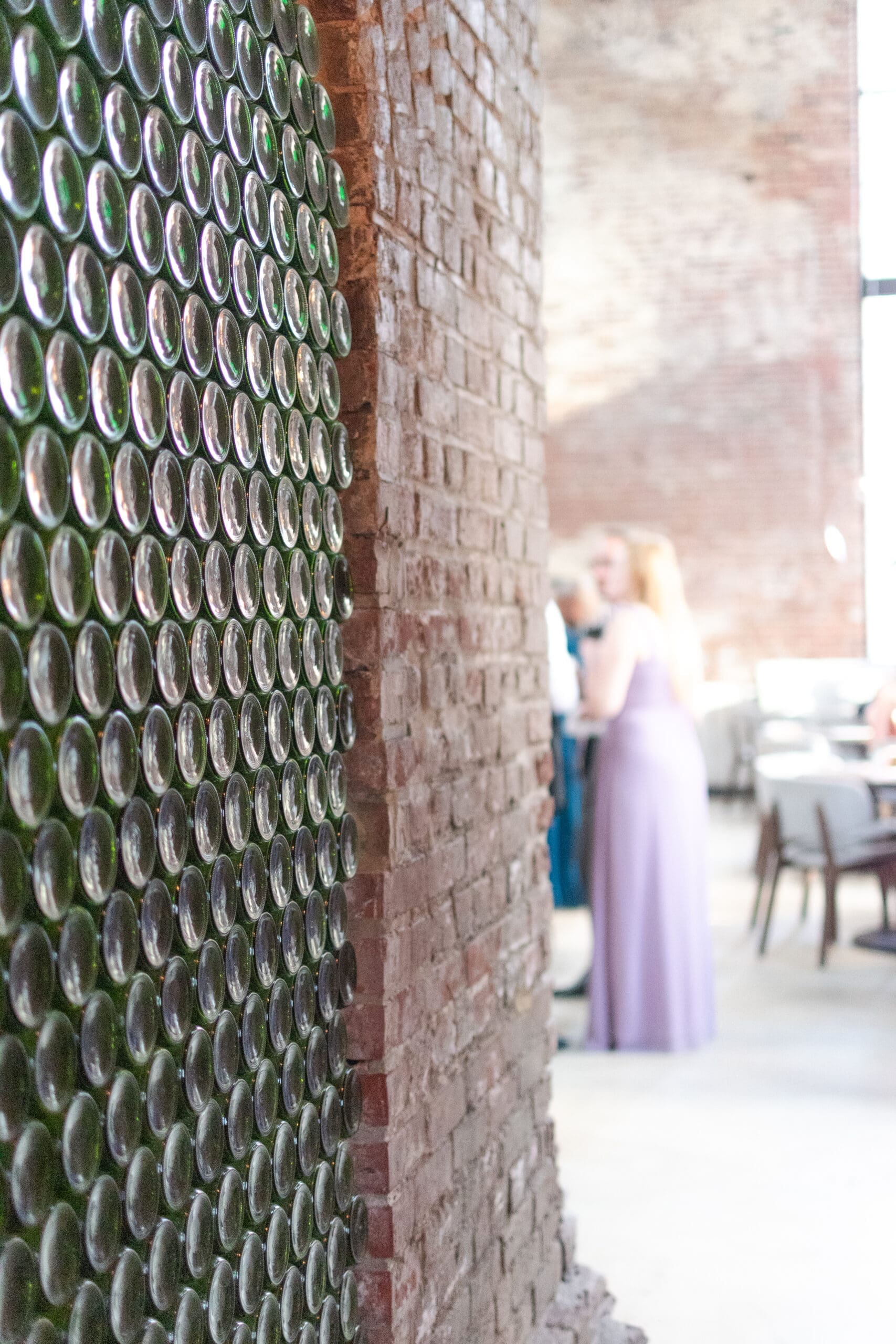 Close-up of a brick wall with bottle bottoms embedded. In the blurred background, two people converse near dining tables, creating a relaxed atmosphere.