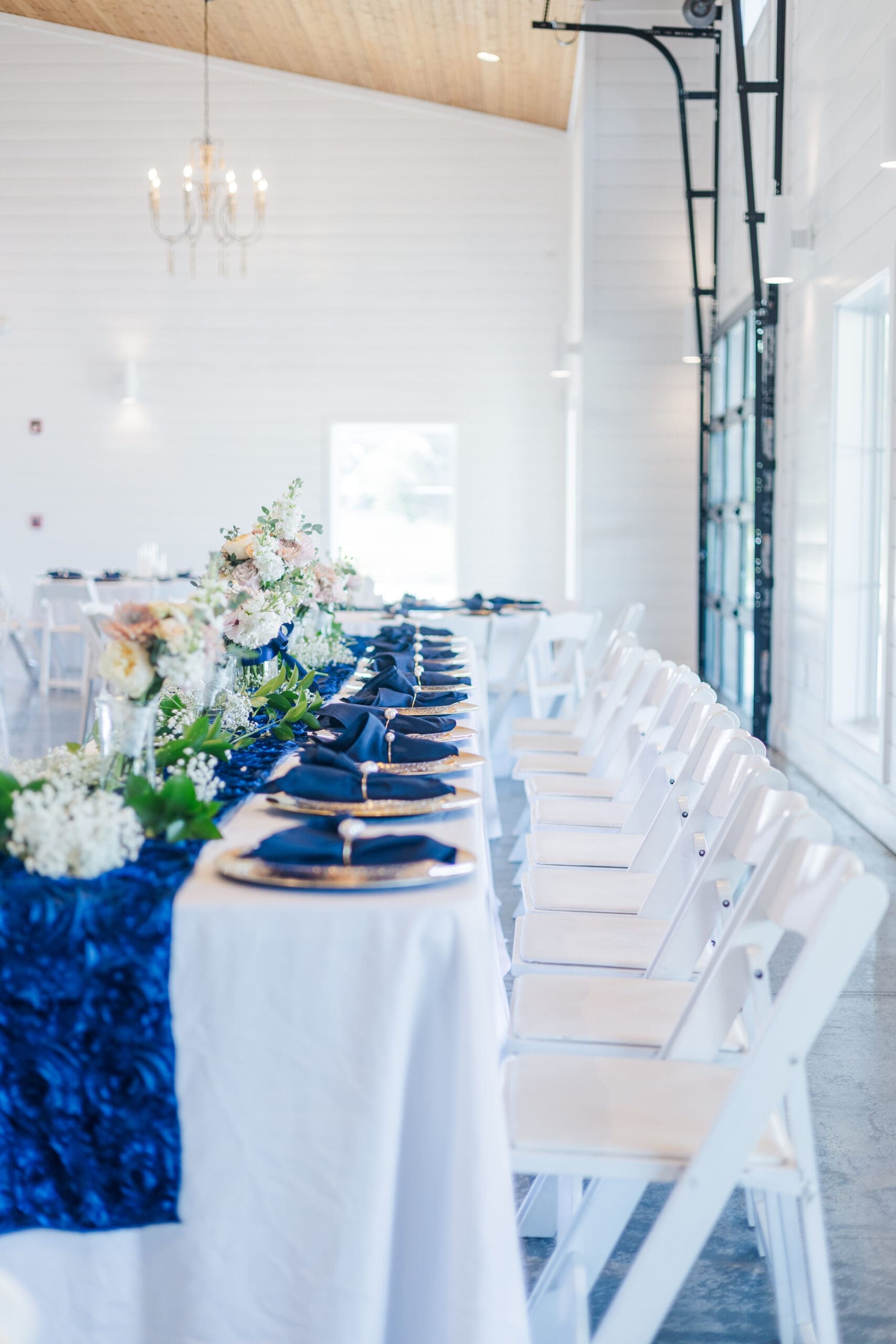 Elegant wedding reception setup with a long table, adorned with blue and white linens, gold chargers, and floral centerpieces. White chairs line the table, set up by their Maryland wedding planner.