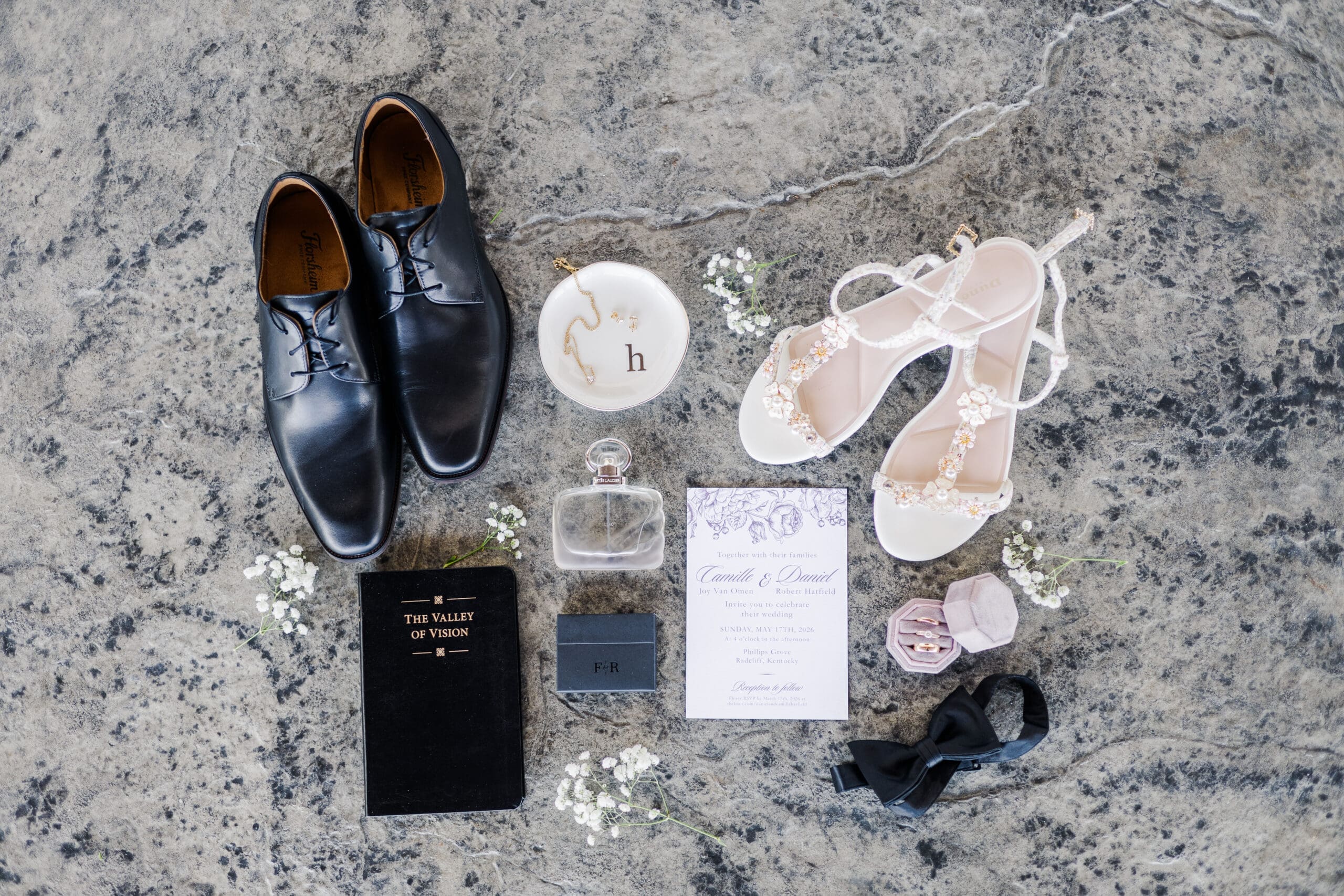 Flat lay of wedding essentials on a textured surface: black dress shoes, white heels, invitation, ring dish, perfume, vow book, and floral elements. Elegant and romantic.