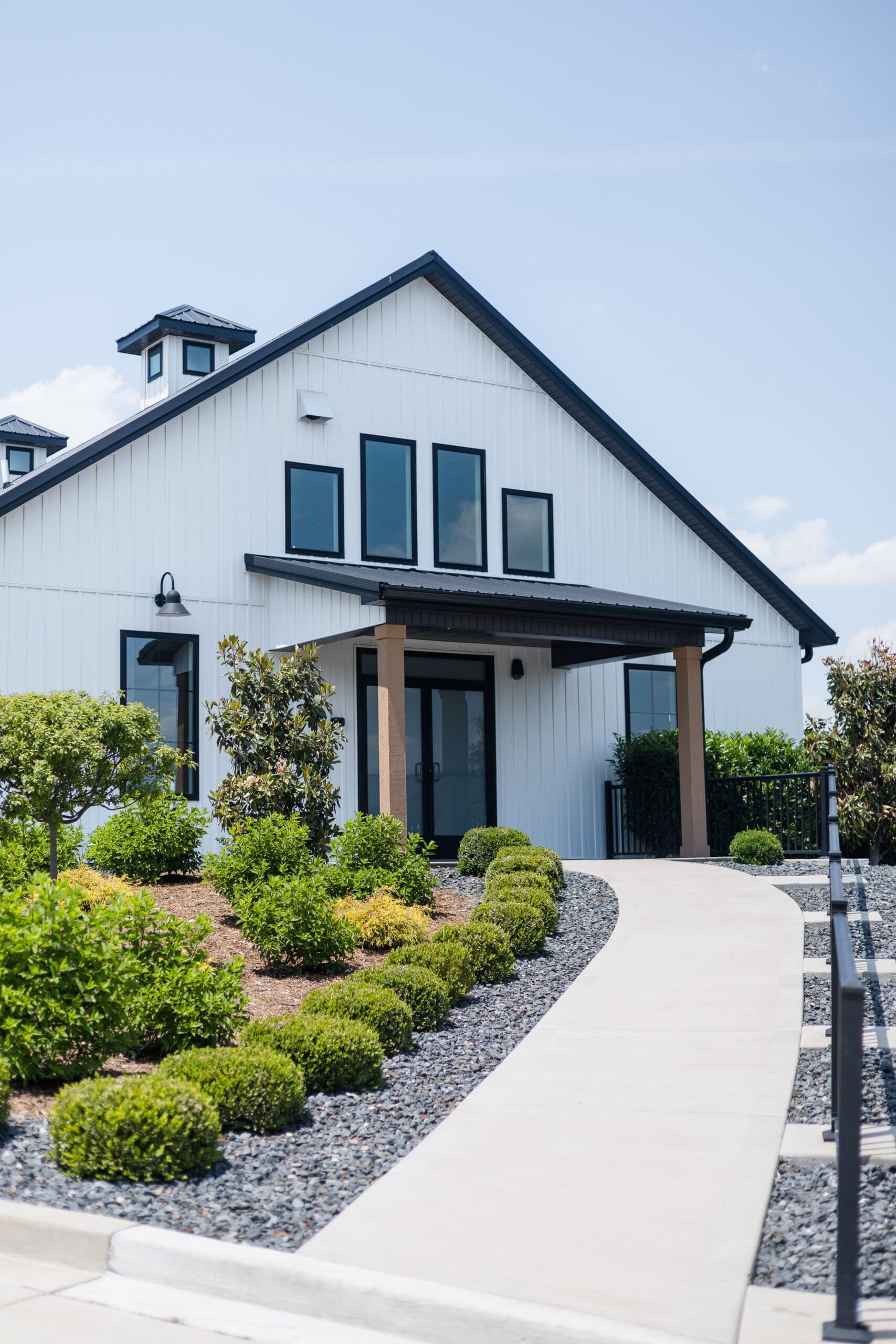 Beautiful white ceremony A-frame building with black and wood accents. Green landscaping and a sidewalk path lead to the building.