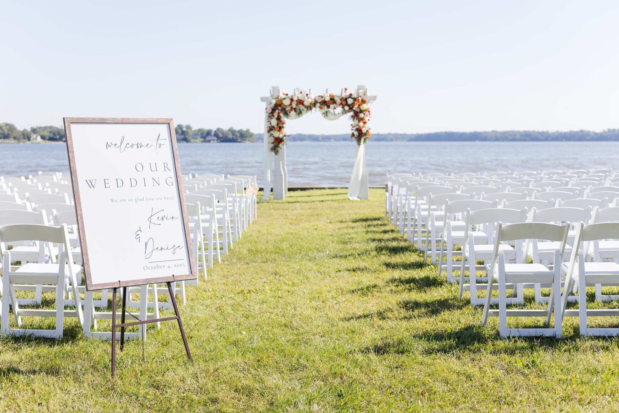 Outdoor wedding setup by a lake, featuring rows of white chairs on green grass, a floral arch, and a welcome sign on an easel, all under a clear blue sky.