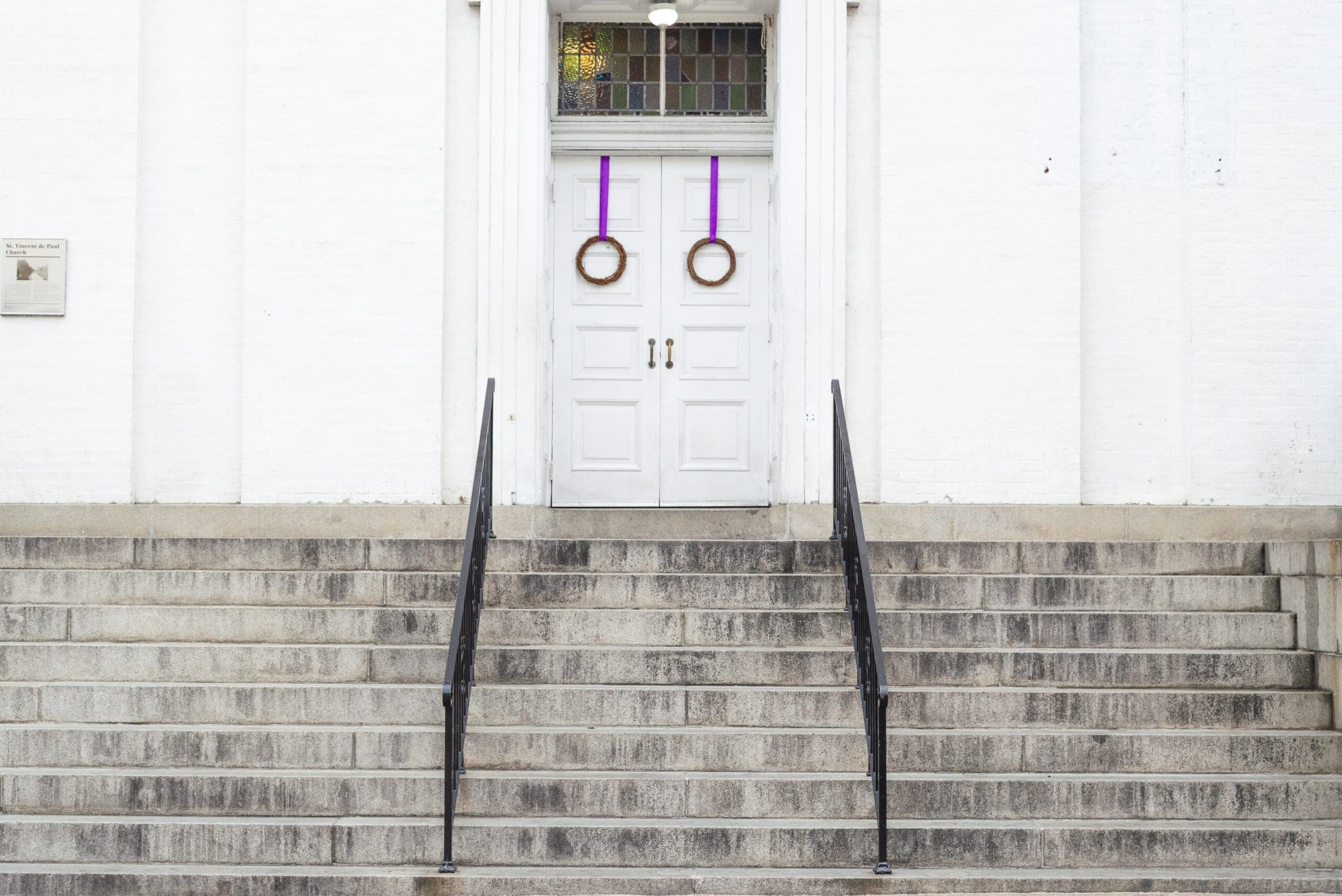 Purple wreaths hang on white doors of a church building. cement stairs lead up to the doors.