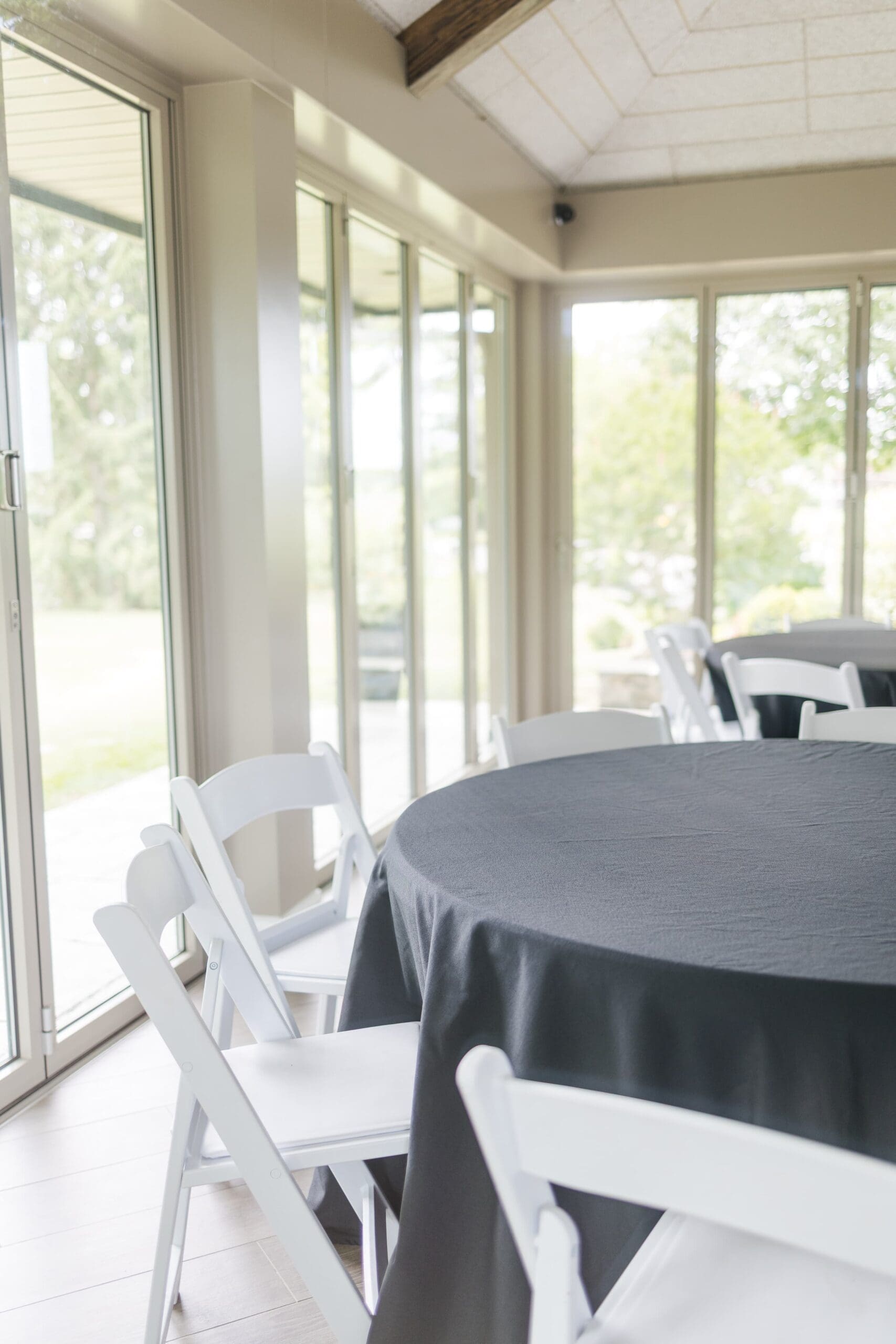 A dining area featuring a round table with a black tablecloth and white chairs, surrounded by large windows offering a view of the outdoors.