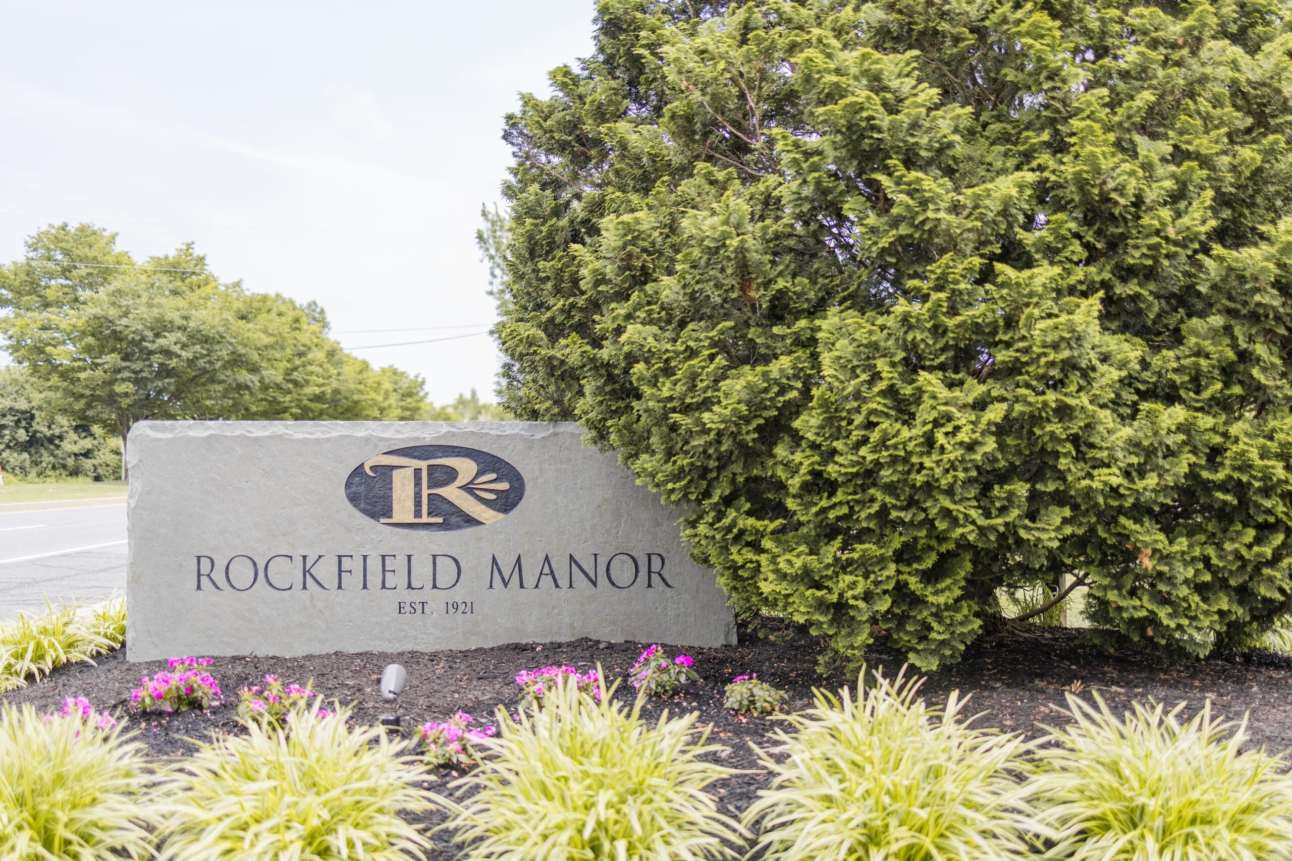 Stone sign reading "Rockfield Manor, Est. 1921" with a decorative "R" logo, surrounded by lush green shrubs and small flowering plants. Bright, serene setting.