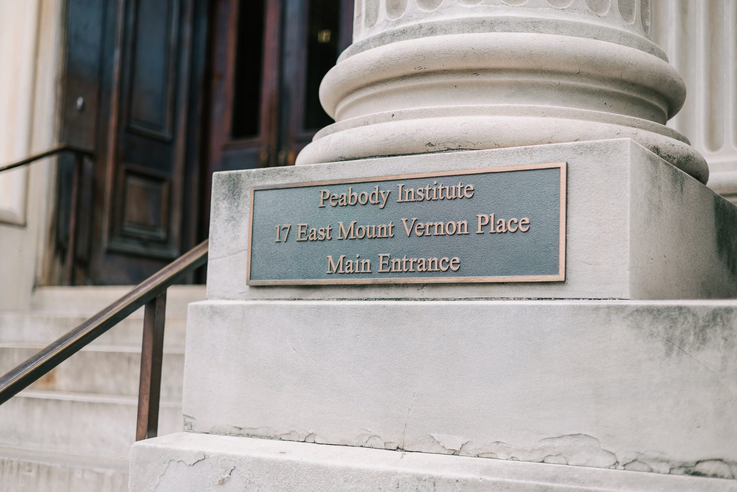 A weathered stone pillar, featuring a metal plaque with "Peabody Institute" written on it stands beside steps leading to a building entrance, evoking a sense of history and importance.