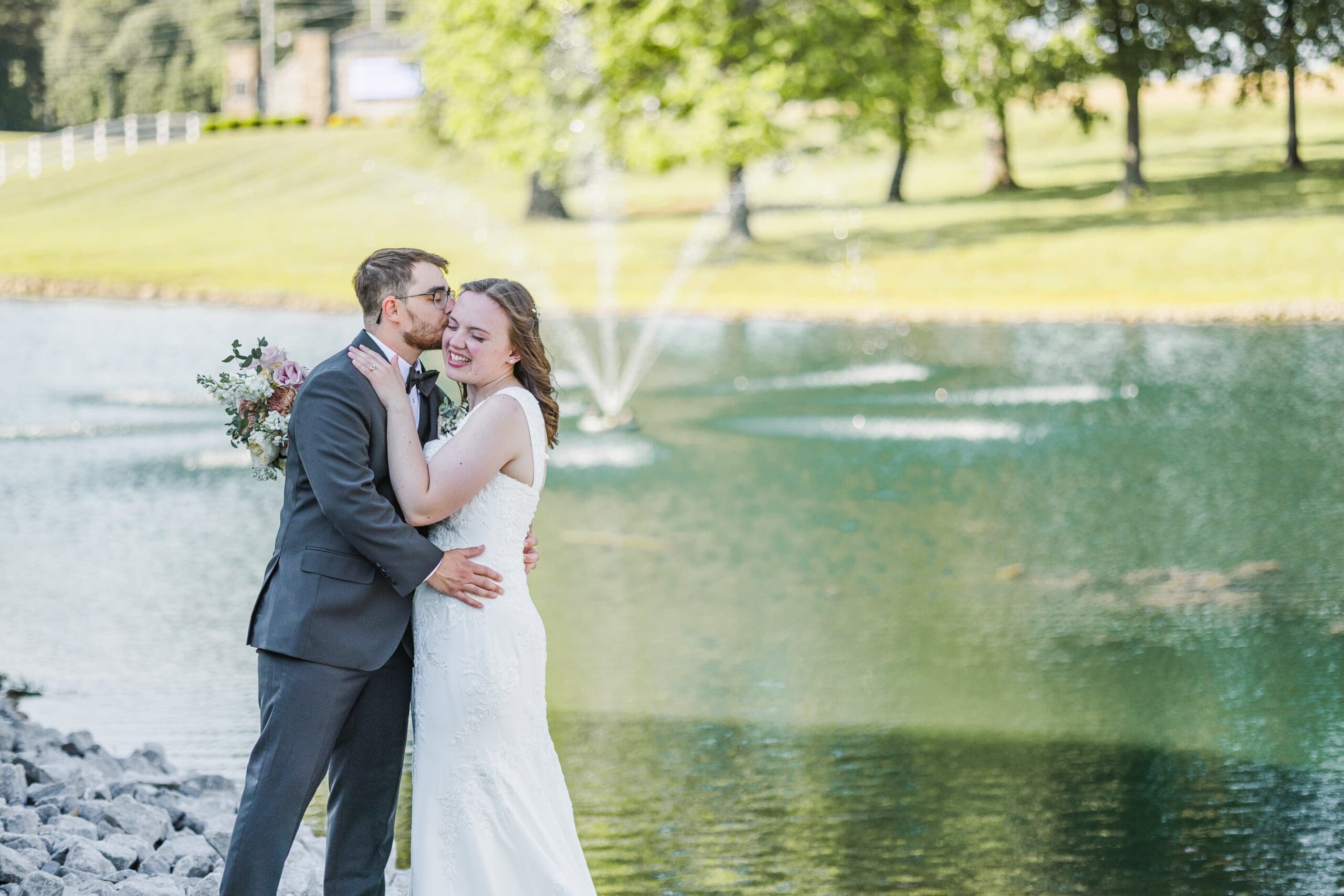 A newlywed couple embraces by a serene pond, with a fountain and lush trees in the background. The groom kisses the bride's cheek, both smiling warmly.