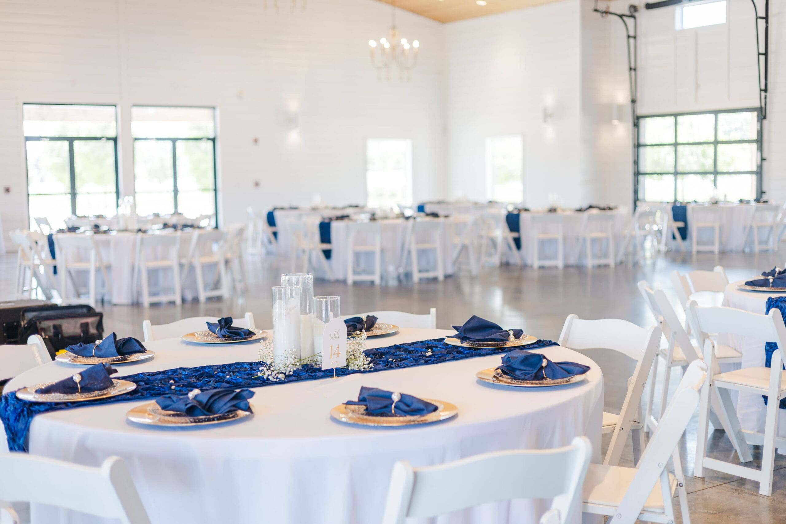 white tableclothed tables line a white room with wood floor. The space is bright and white. Blue tablerunners line each table.