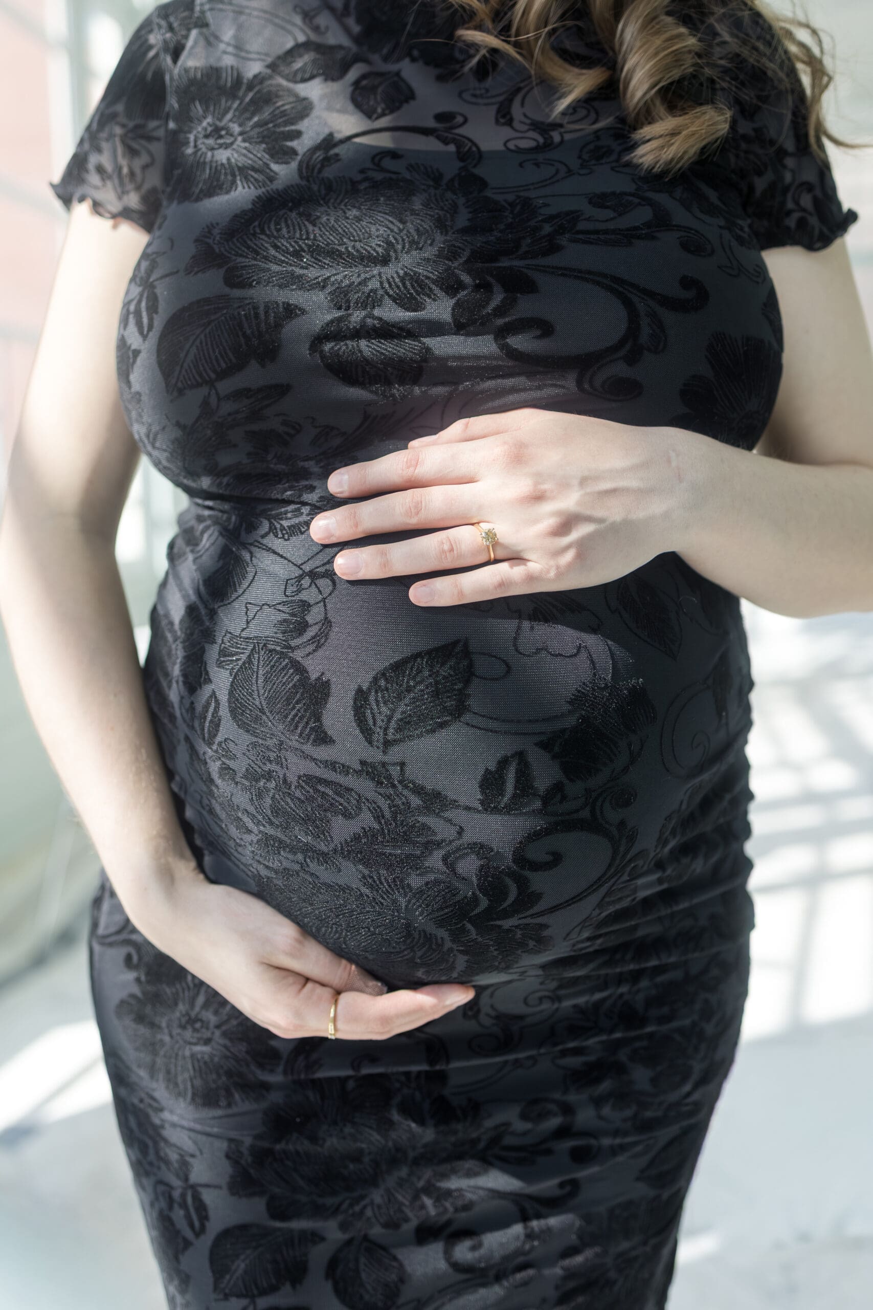 Pregnant woman in a black lace dress gently holding her belly with both hands, conveying warmth and anticipation, in soft natural light.