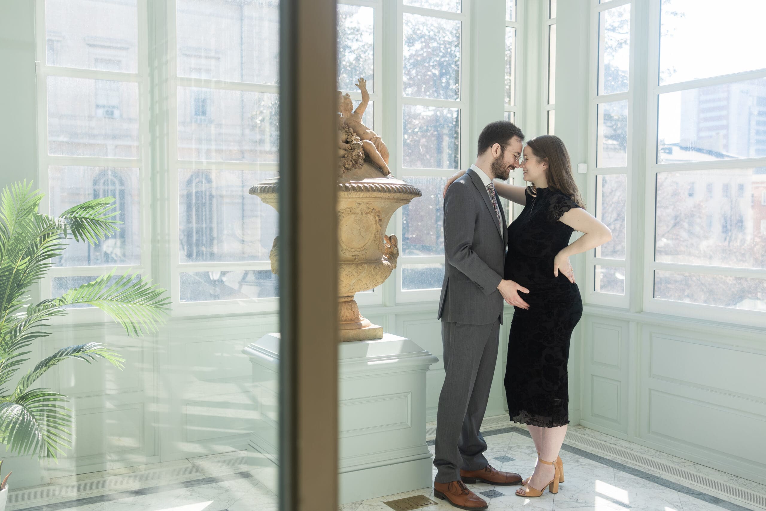 Pregnant woman in black lace dress leans against her husband in gray suit as he holds her belly in a sunlit room. They nuzzle together close, anticipating the arrival of their baby.