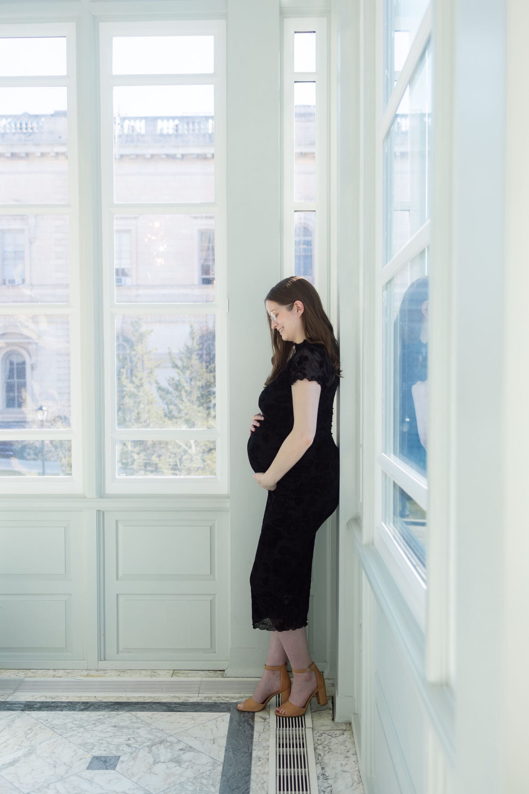 Pregnant woman in a black lace dress gently holding her belly leaning against the wall as she looks down at her belly, conveying warmth and anticipation, in soft natural light.