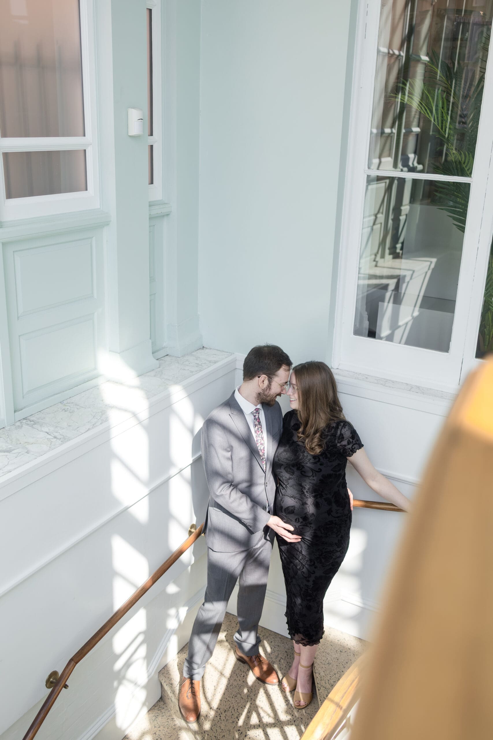 Pregnant woman in black lace dress holds her forehead to her husband in a gray suit. They stand in a sunlit stairwell nuzzling close to one another.