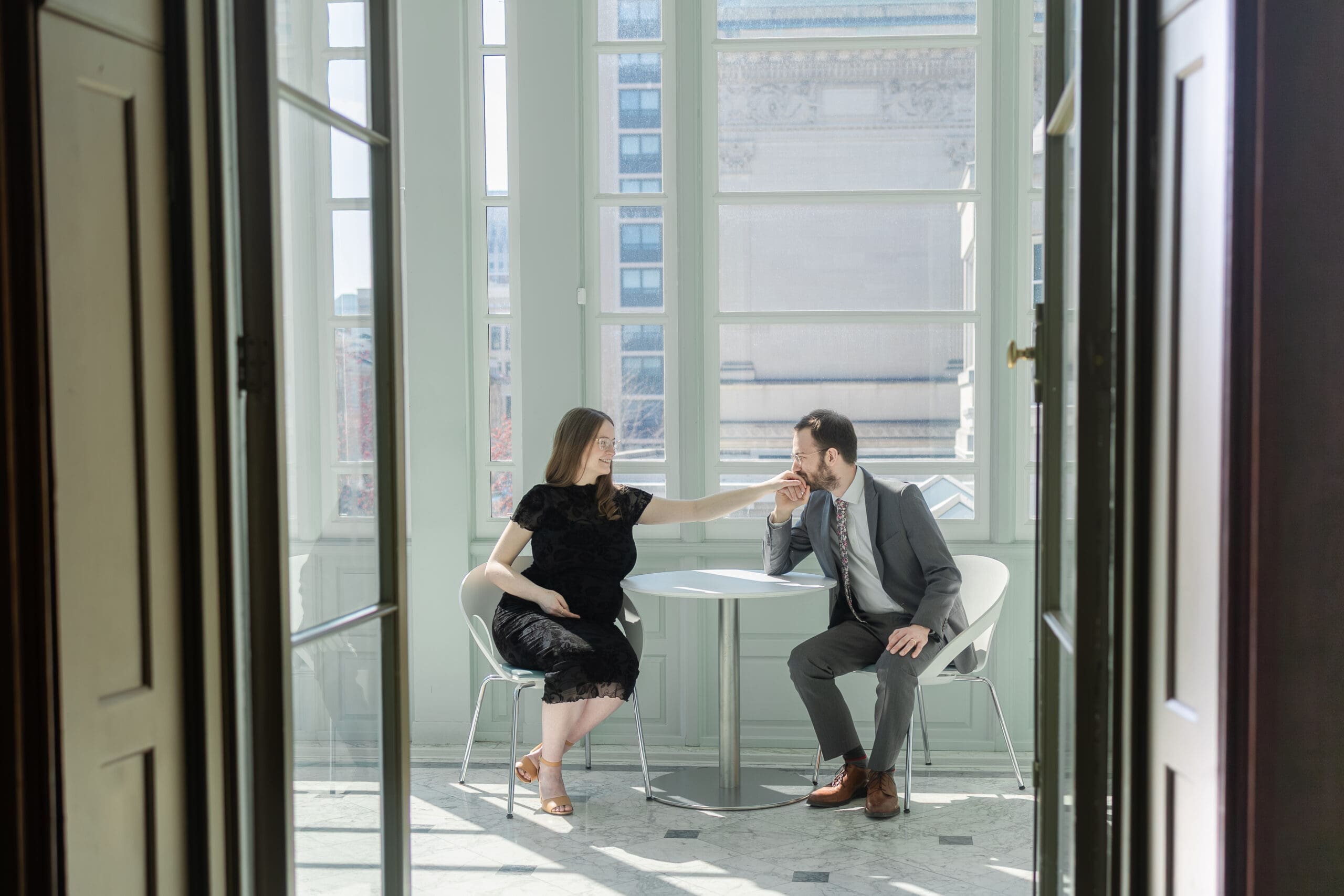 Pregnant woman in a black lace dress holding out her hand as her husband in a gray suit kisses it gently. They sit in a sunlit room leading from brown doors.