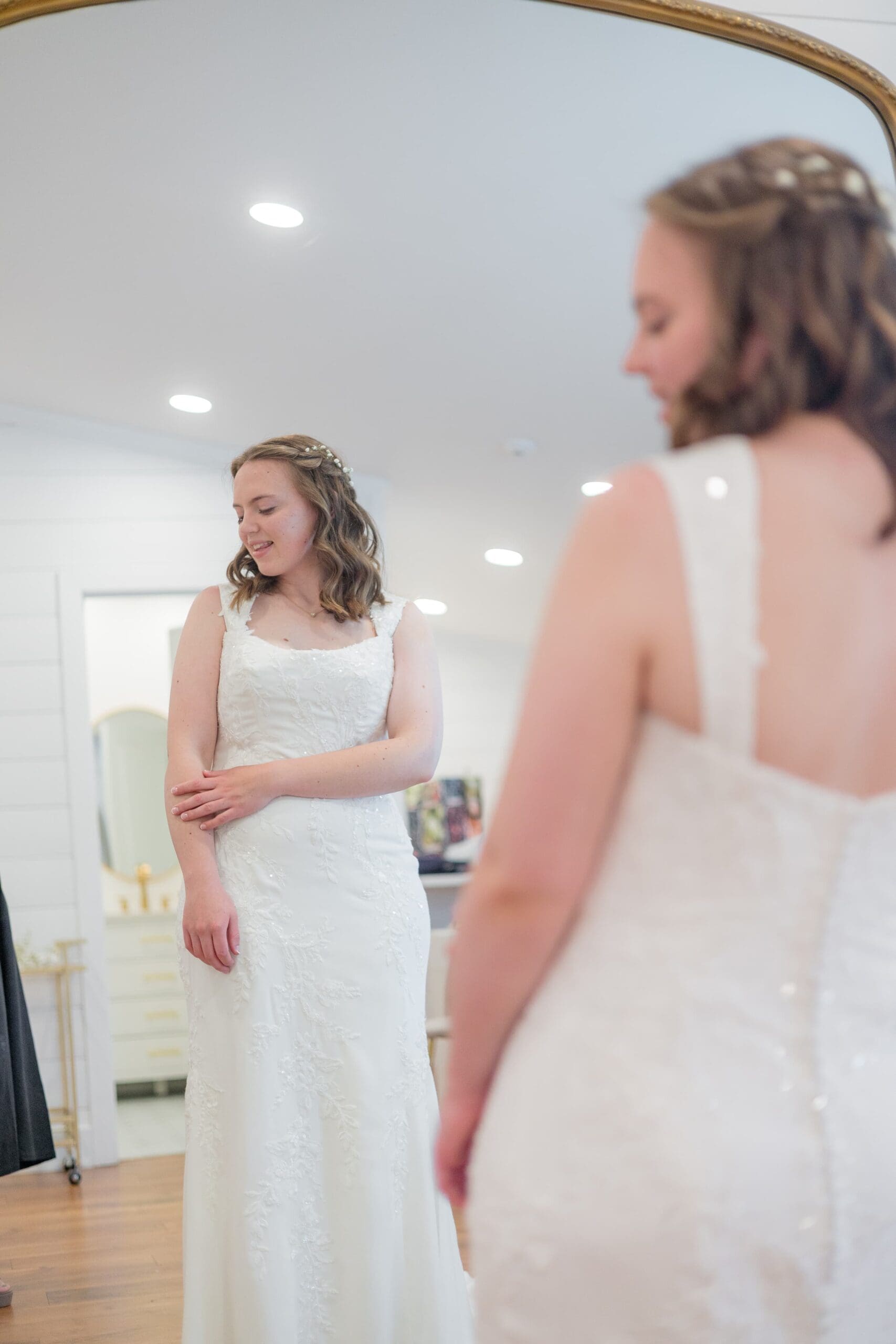 A bride in her wedding dress stands in the mirror, looking down softly. 