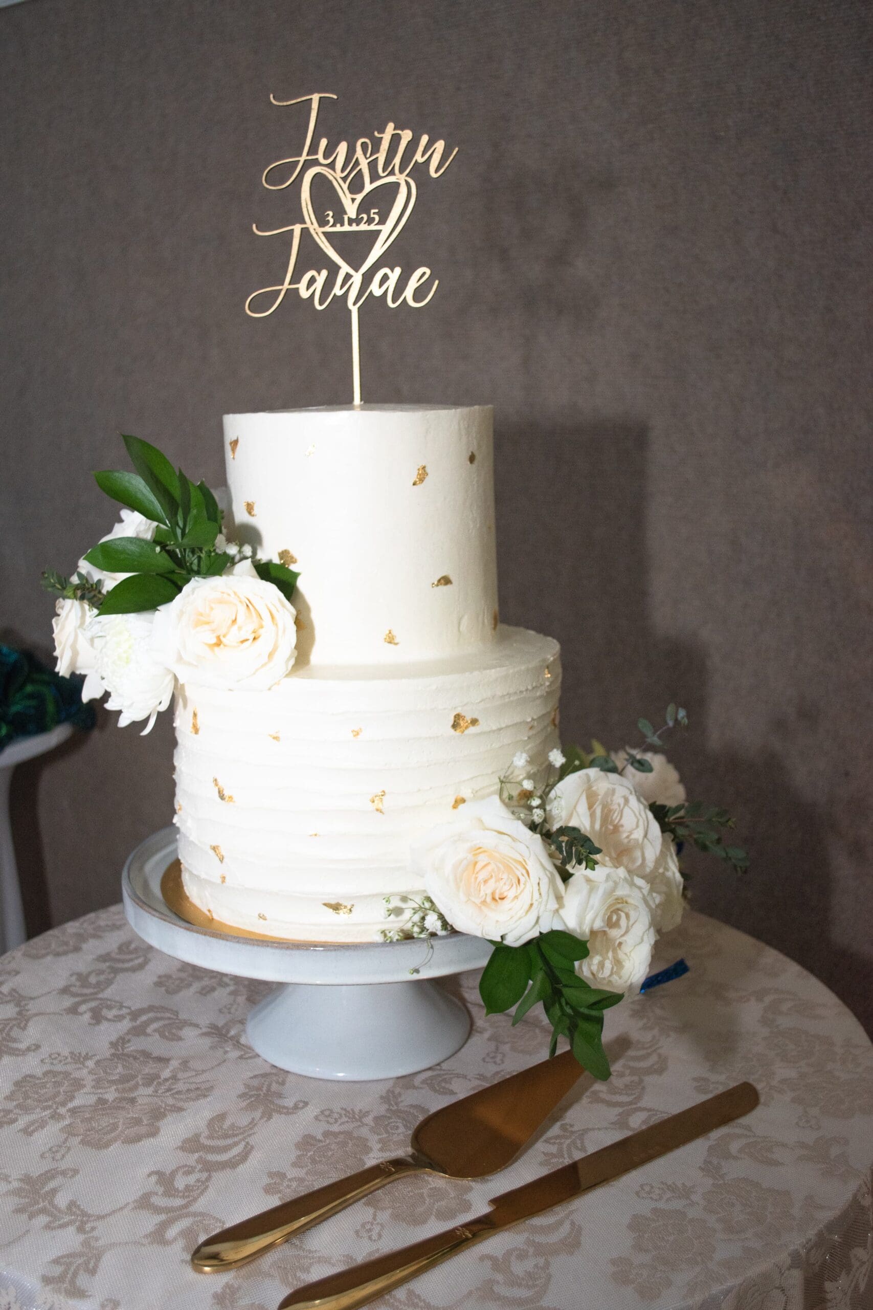 Flash edited wedding cake on a stand. The cake has gold pieces on it with a cake topper that says, "Justin & Janae" with a heart.