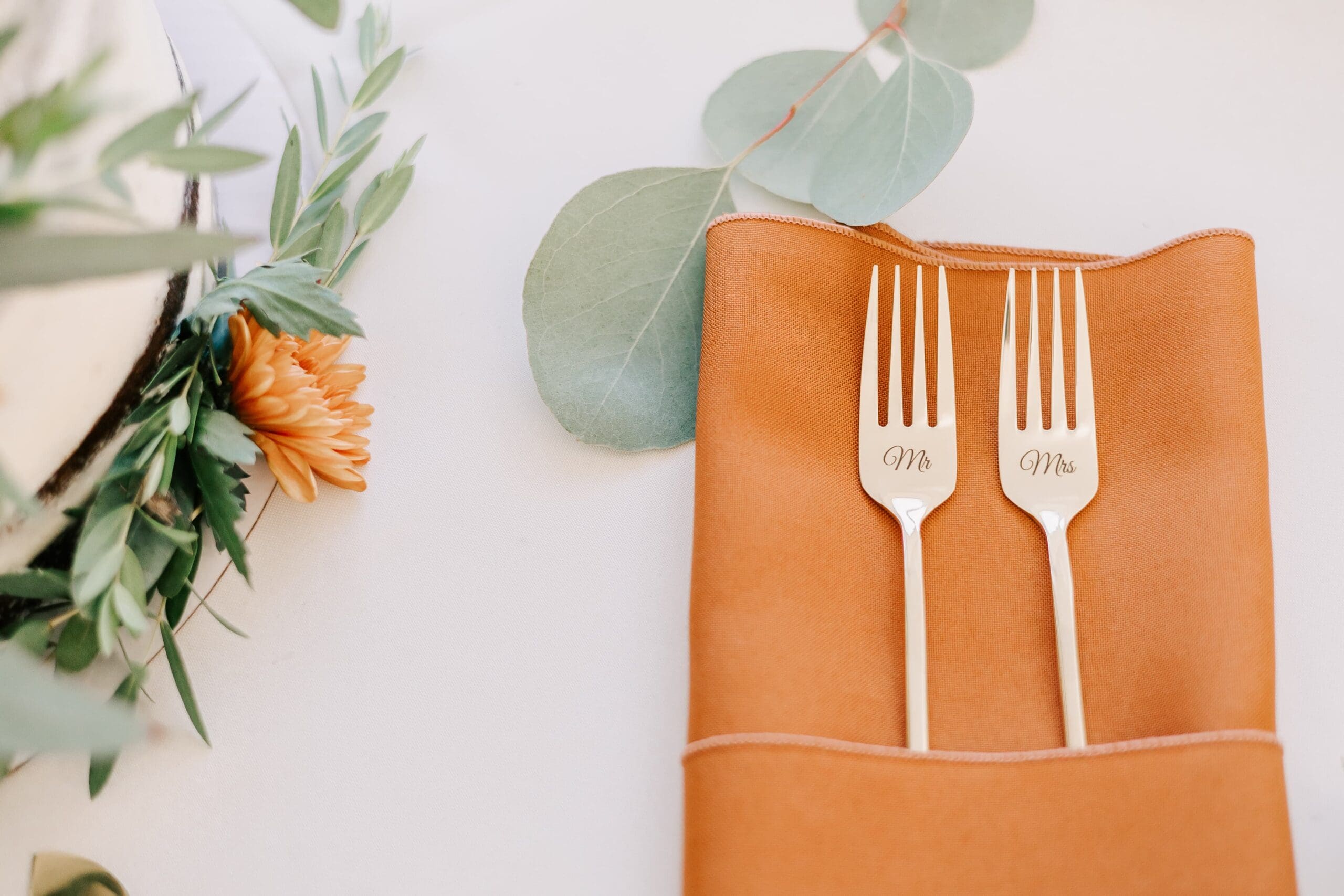 Two elegant forks rest on an orange napkin, placed on a white table. Eucalyptus leaves and a small orange flower add a fresh, decorative touch.