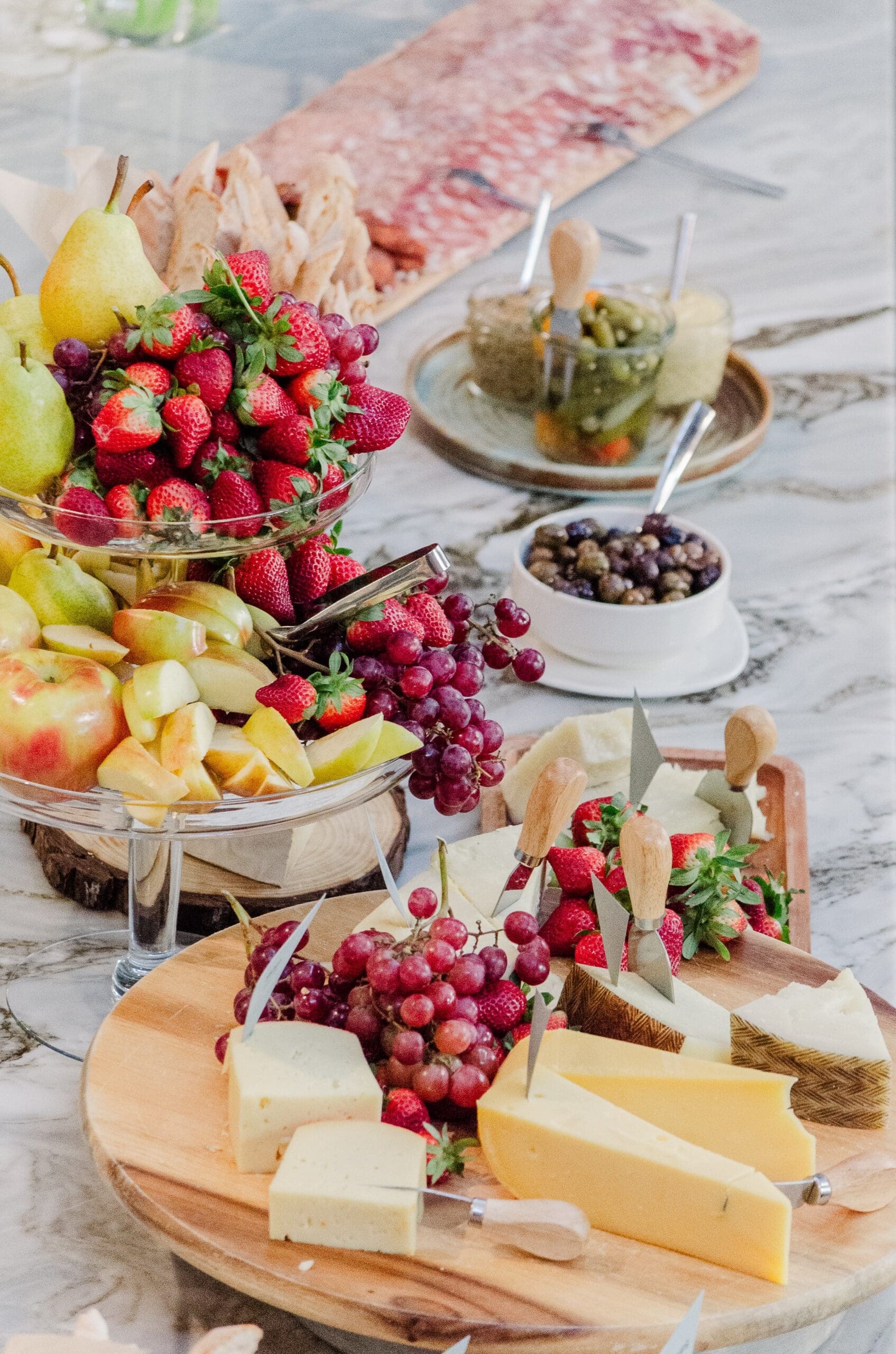 A colorful spread of fresh fruits, including strawberries, grapes, and melon, on tiered trays alongside a variety of cheeses, creating an elegant and inviting display.