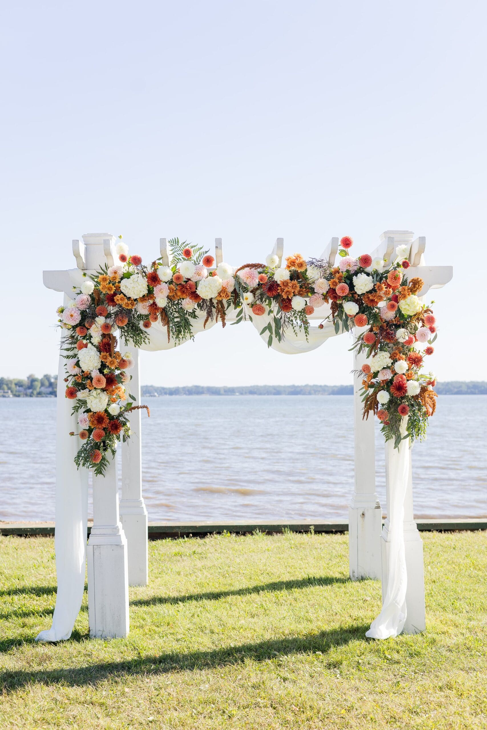 Outdoor wedding arch setup by a lake, featuring green grass, a floral arch with orange white and green, all under a clear blue sky.