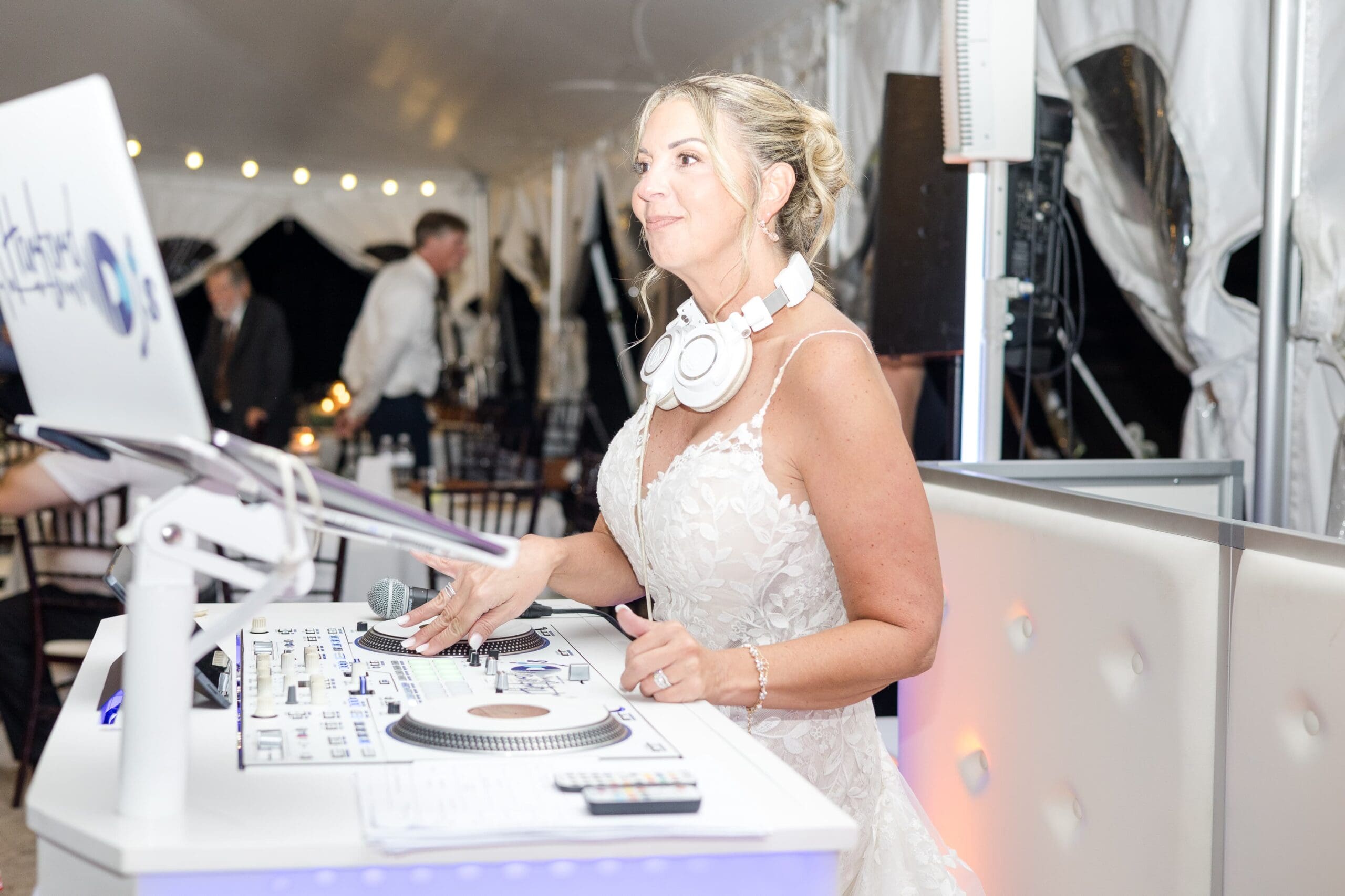 Bride behind the DJ booth. She is in a white tent reception space with DJ headphones around her neck, moving the discs on the sound board with a smile on her face.