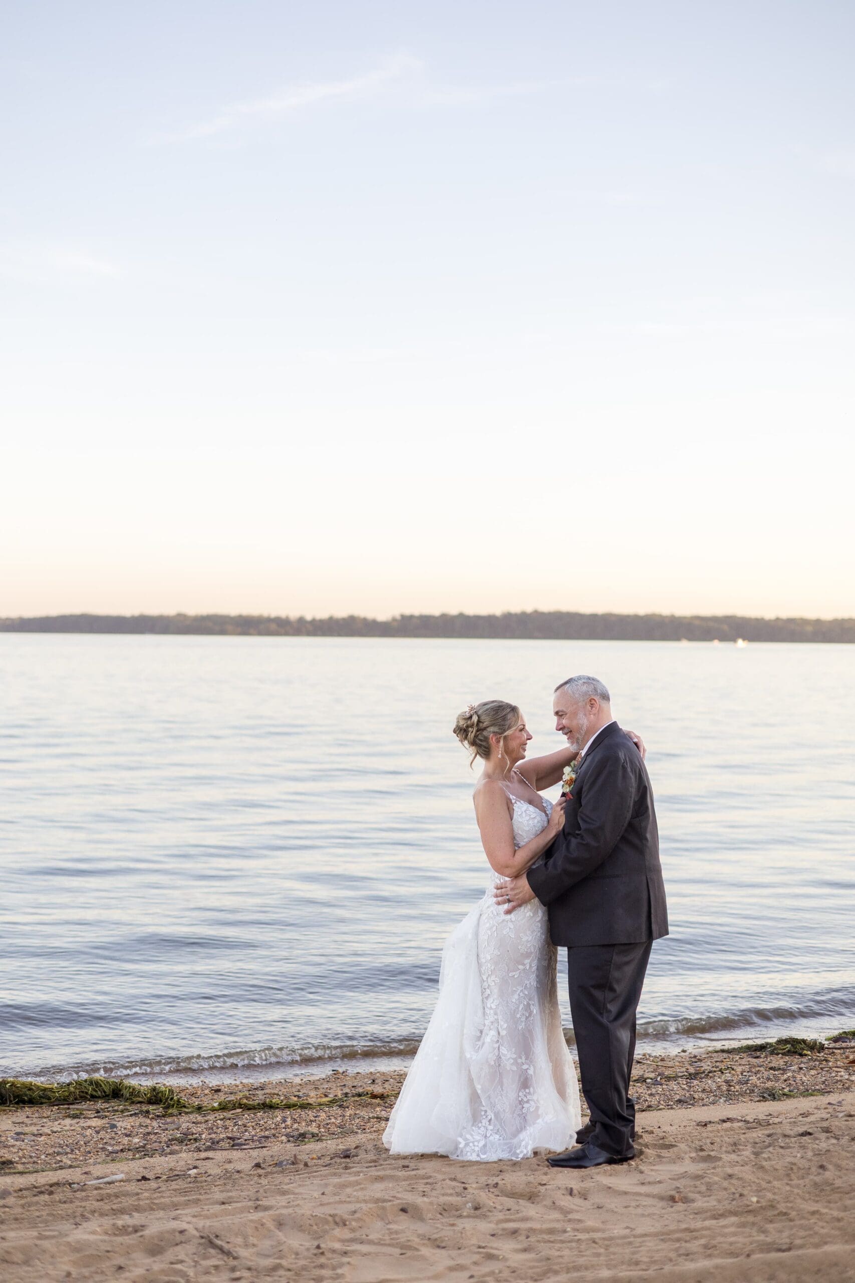 A newlywed couple embraces joyfully on a sandy beach. The woman wears a bridal gown, with a serene backdrop.