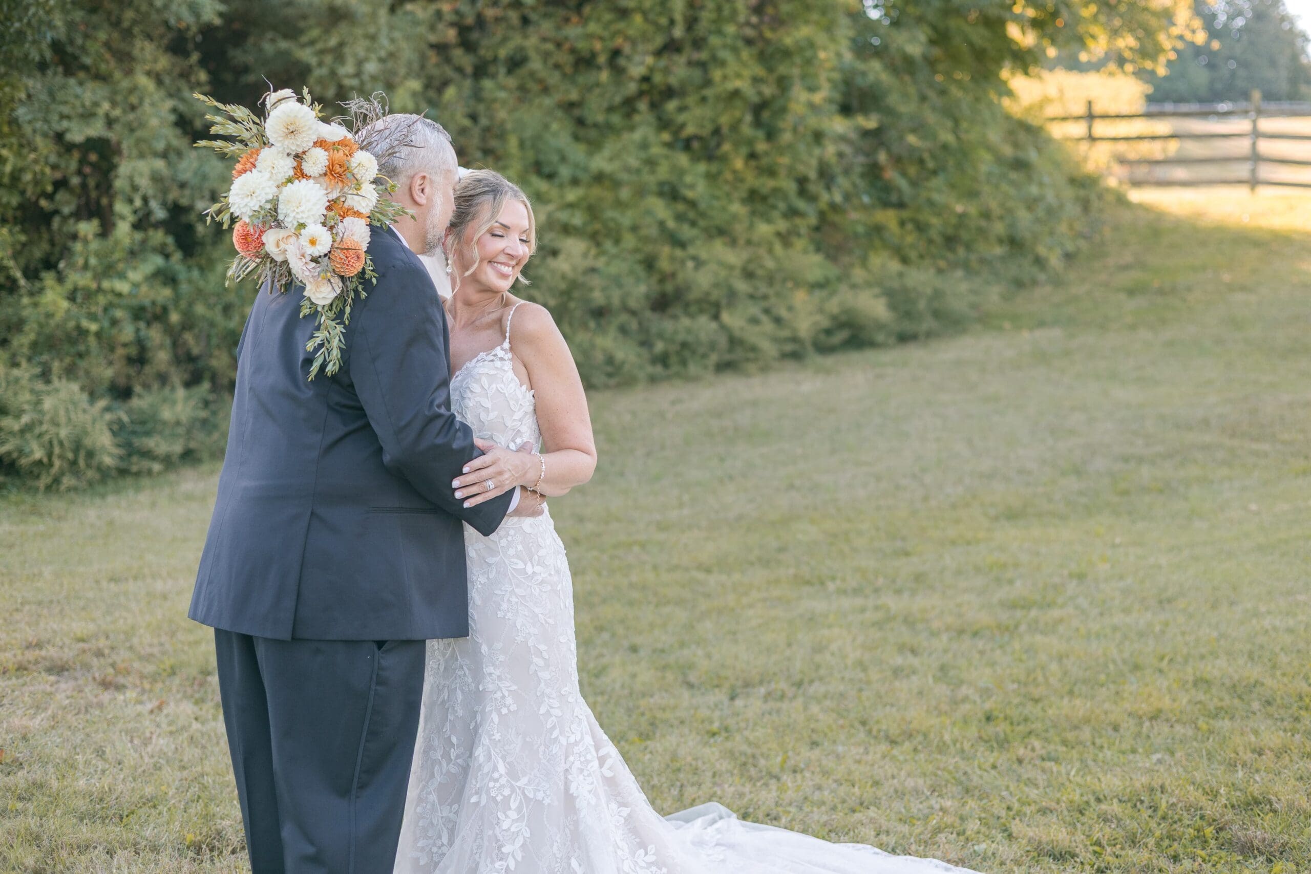 A newlywed couple embraces joyfully in a grassy field. The woman wears a bridal gown, holding a bouquet of orange and white flowers, with a serene backdrop.