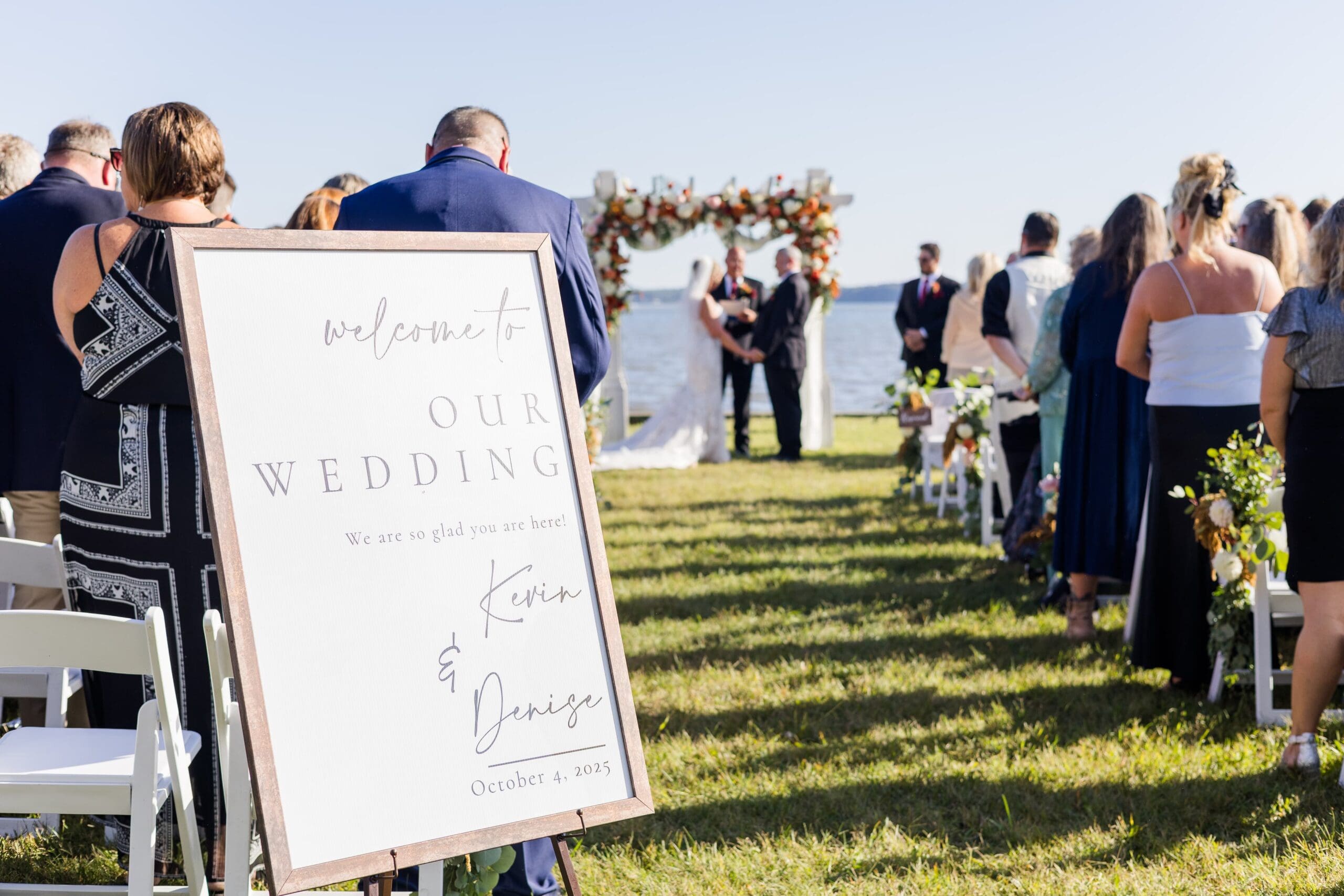 Outdoor wedding setup by a lake, featuring rows of white chairs on green grass, a floral arch, and a welcome sign on an easel, all under a clear blue sky. Bride and groom are blurred in the background at the altar with all of the guests standing at their chairs.