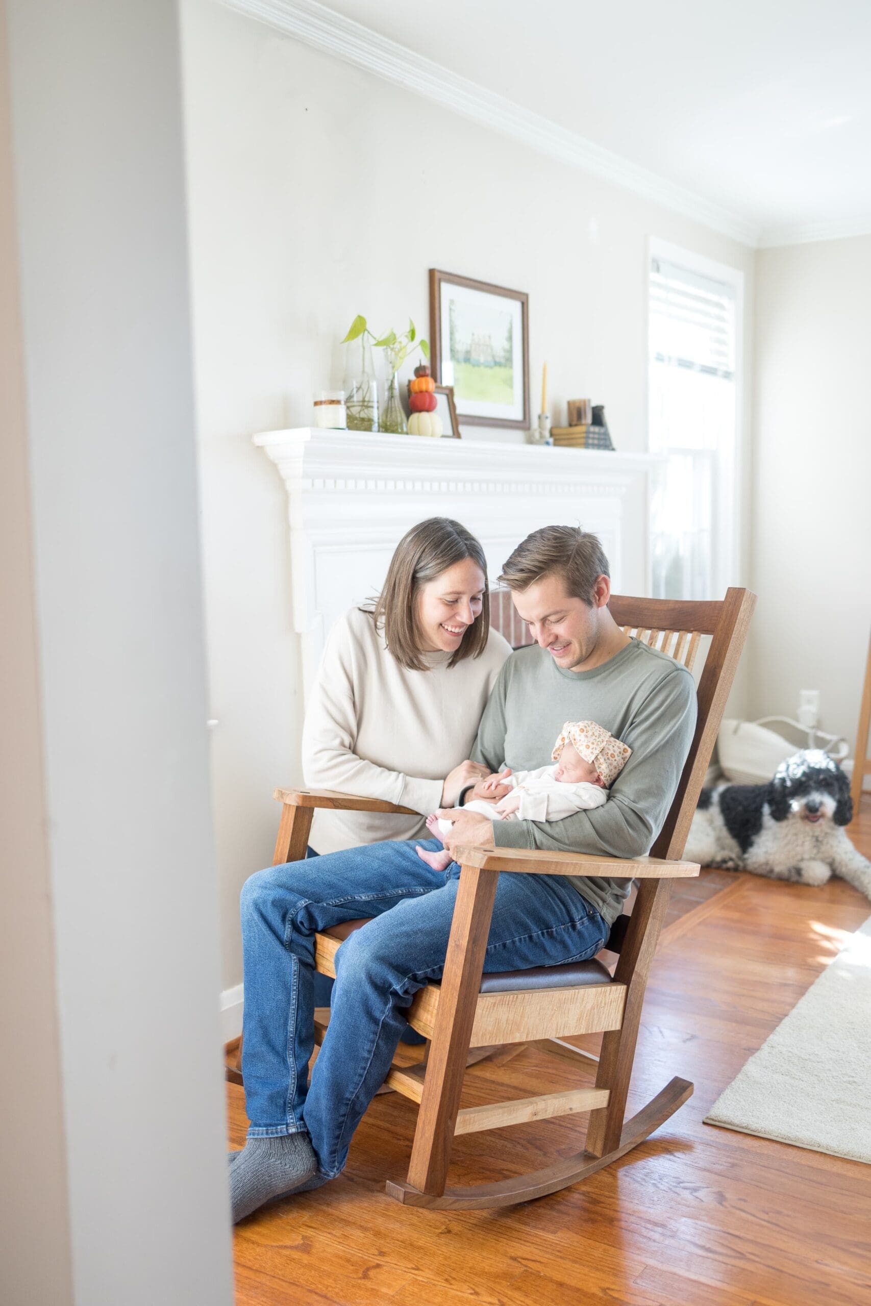 husband and wife nuzzle close and smile as the dad sits on a rocking chair holding their newborn baby