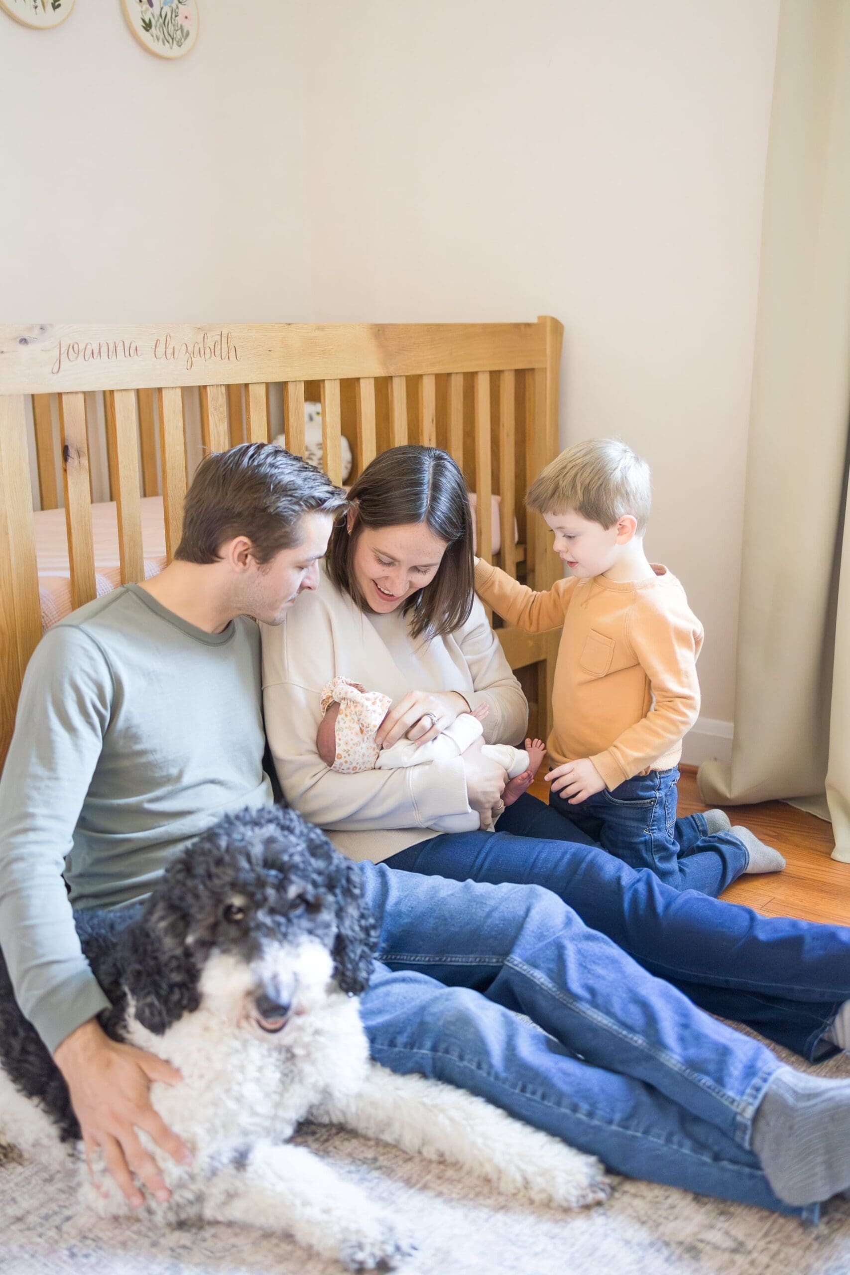 Family sits on the floor in their newborn nursery as they all stare at the newborn while the mom holds her. The dog is there too.