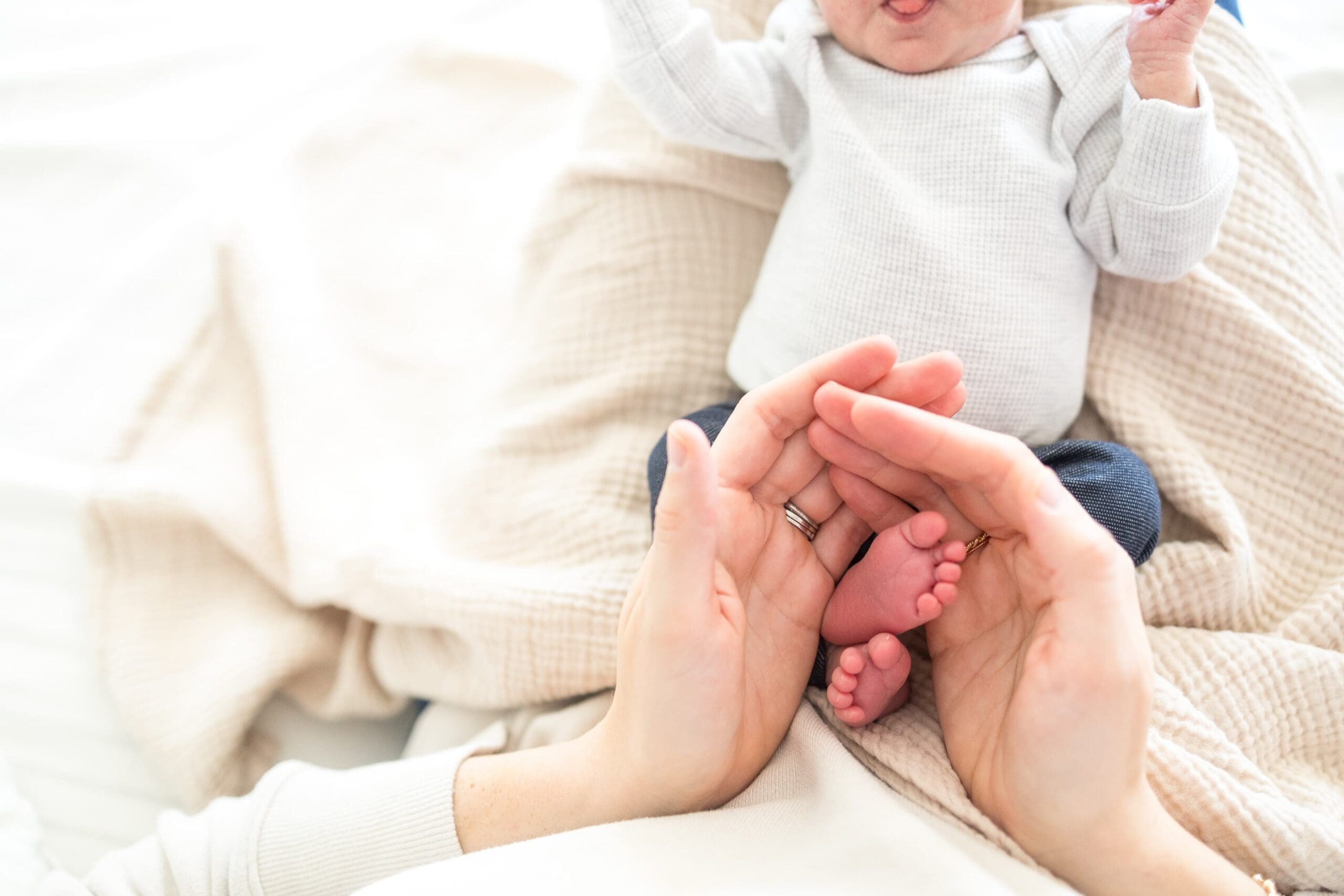 A close-up of an adult gently cradling a baby's tiny feet with both hands. The scene conveys warmth and tenderness, set against a soft, neutral background.