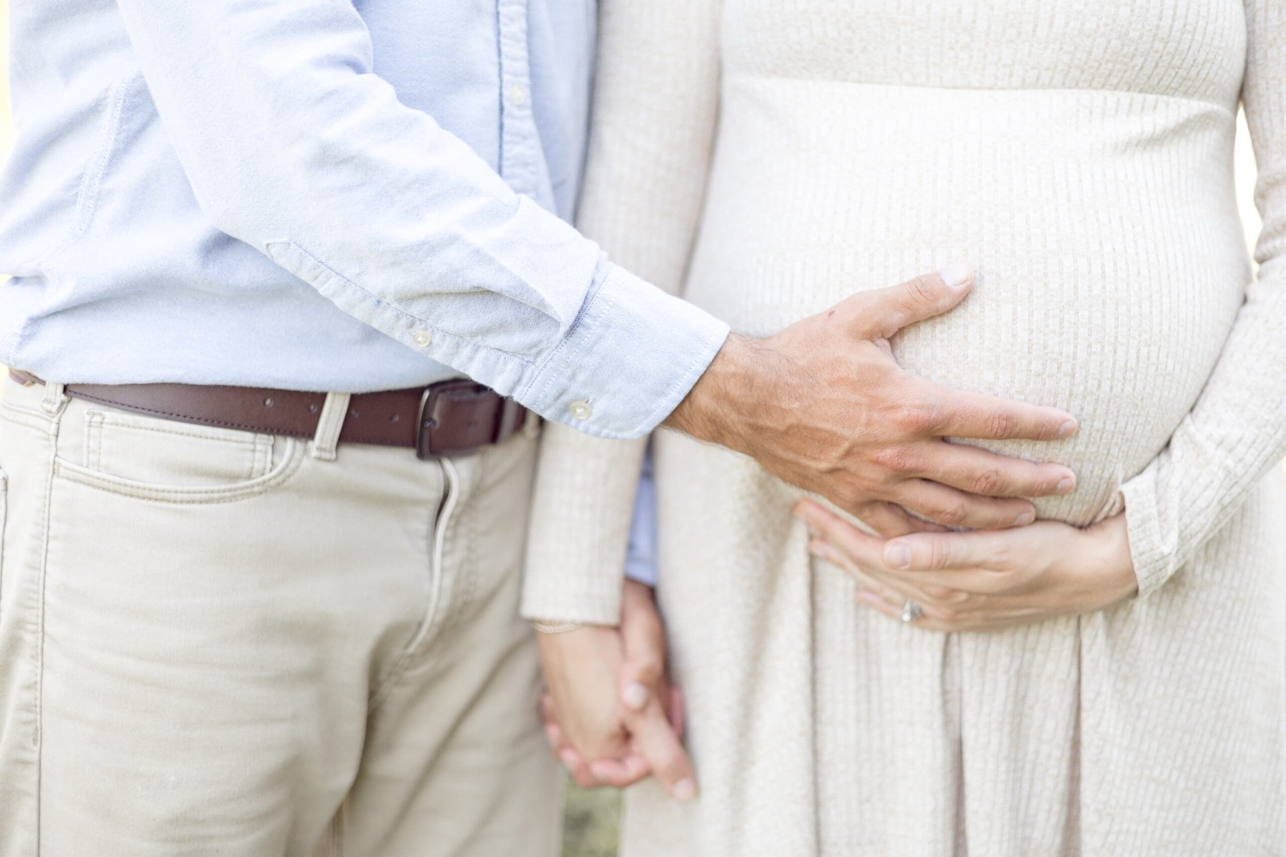 A couple stands close, the man gently placing his hand on the woman's pregnant belly. They are holding hands, conveying a sense of love and anticipation.
