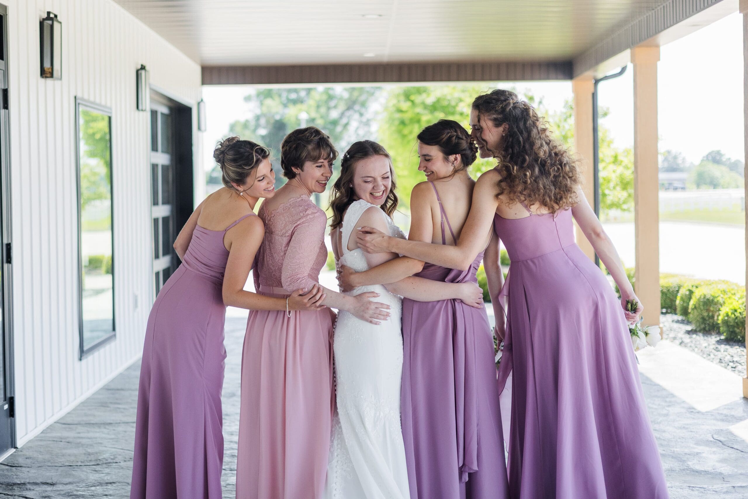 A joyful bridesmaid party poses on a covered patio. They're backs are towards the camera as they get close and hug the bride. Everyone is smiling