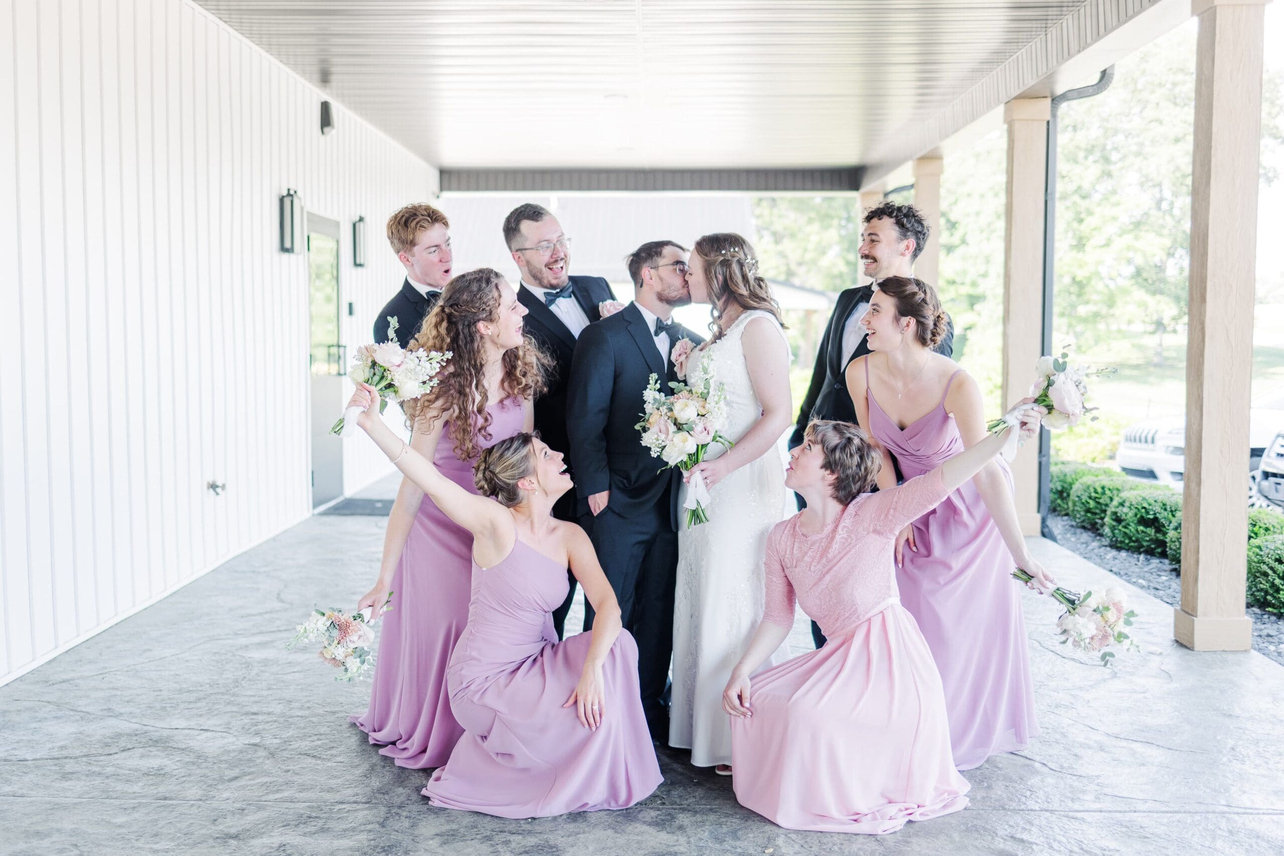 A joyful wedding party poses on a covered patio. The bride and groom kiss in the center, surrounded by bridesmaids in pink dresses and groomsmen in black suits.