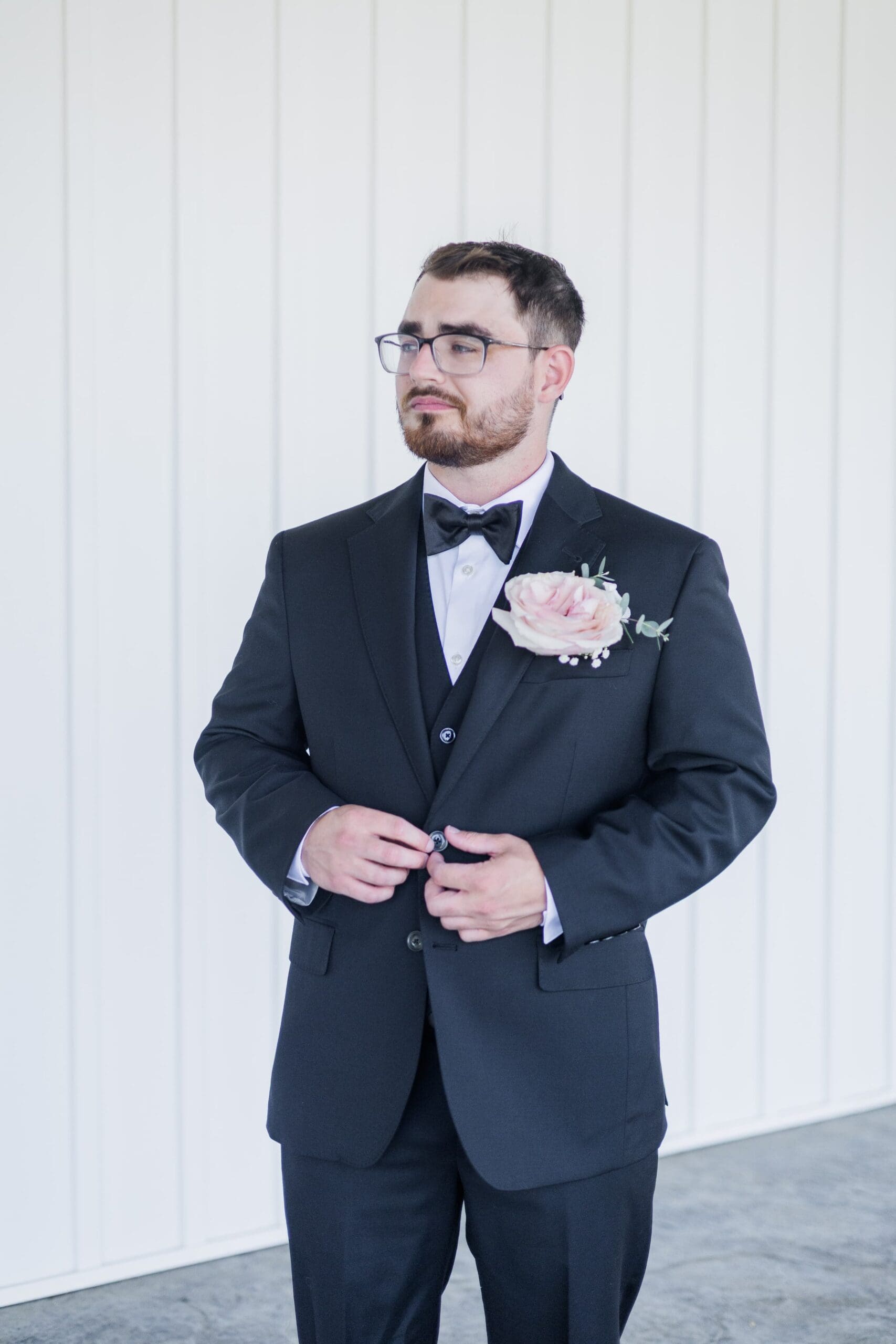 A groom in a black suit looks off into the distance and buttons his suit jacket.