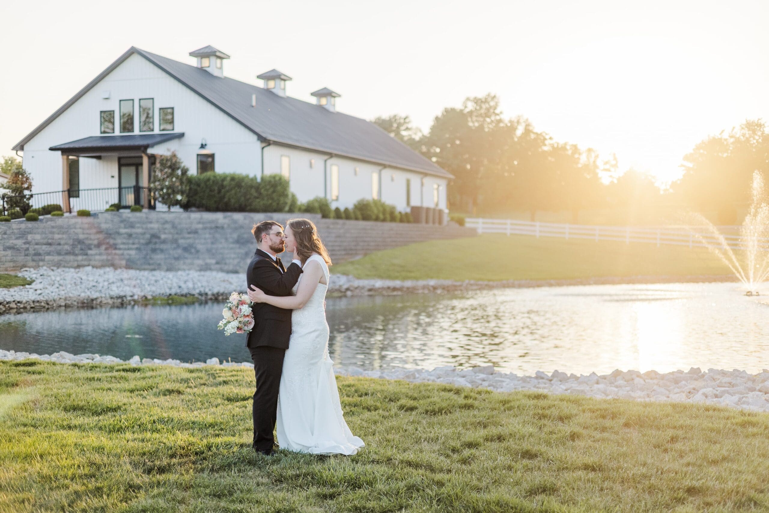 A couple embraces in wedding attire by a tranquil pond at sunset. A white barn stands in the background, exuding a serene and romantic ambiance.