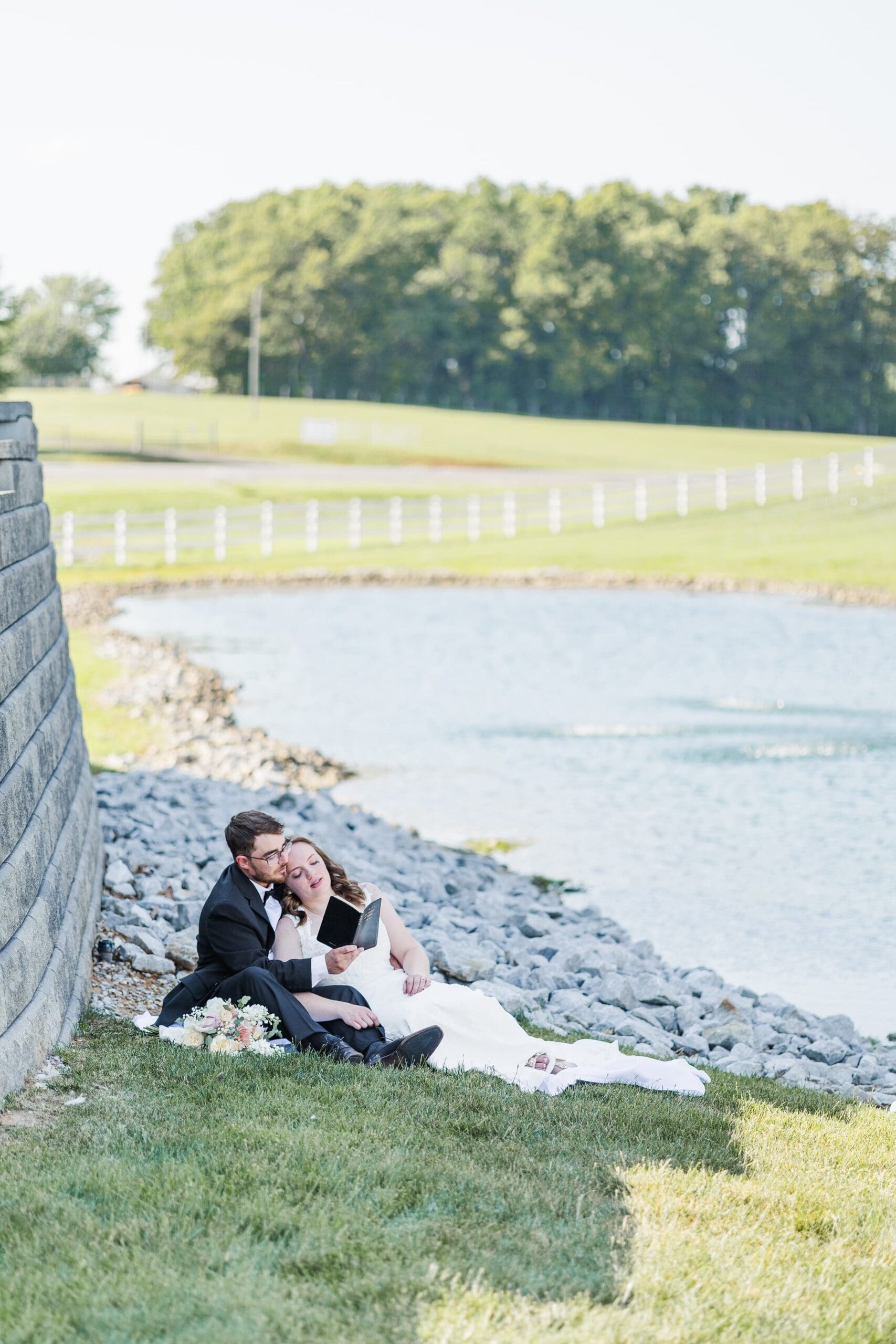 bride in white dress and groom in black tuxedo sit by a lakefront reading a black book on a white sheet in the grass. they are sitting and the bride leans into the groom's shoulder