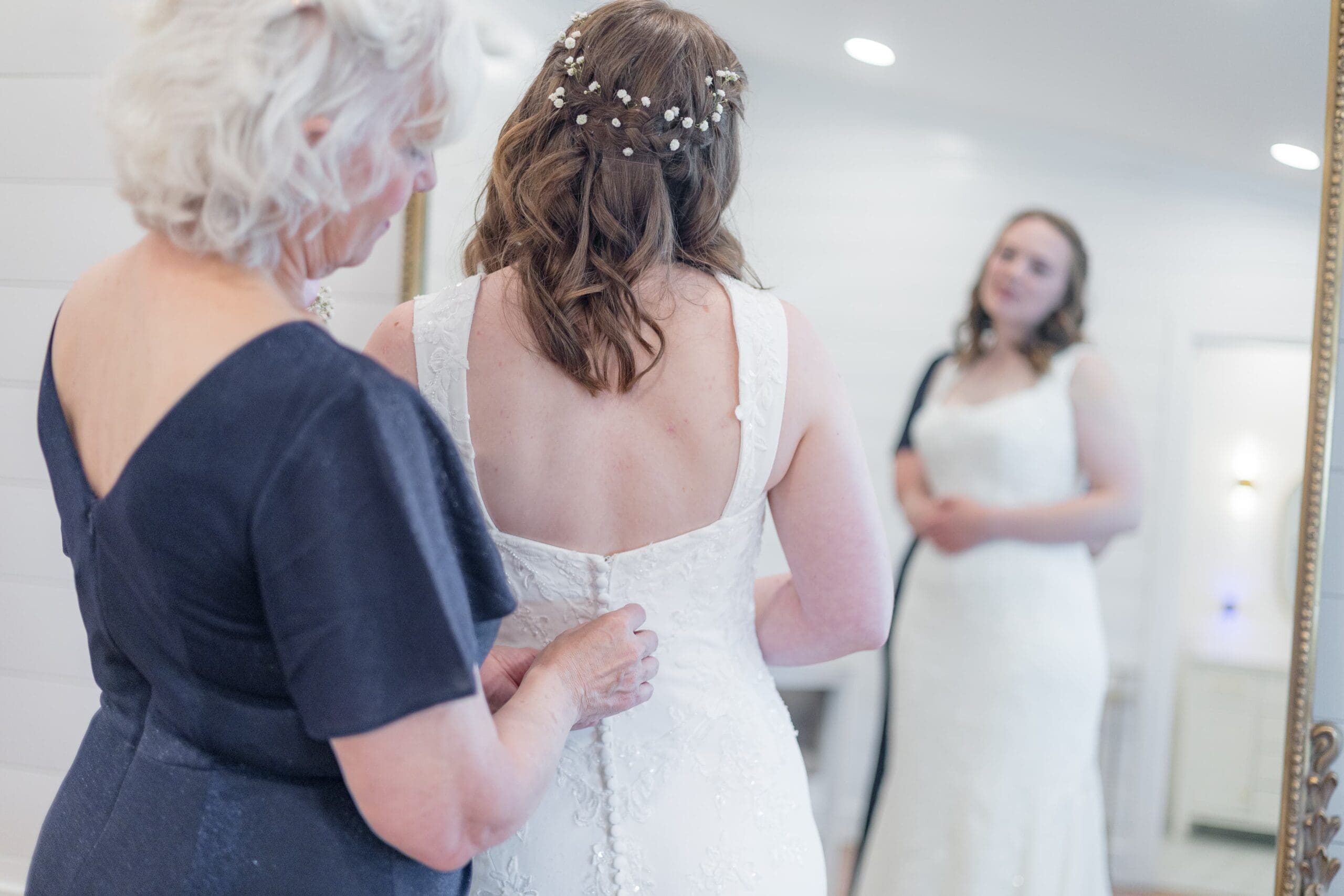 mother in blue formal dress helps button the back of bride's white dress. They stand in front of a mirror in a white room.
