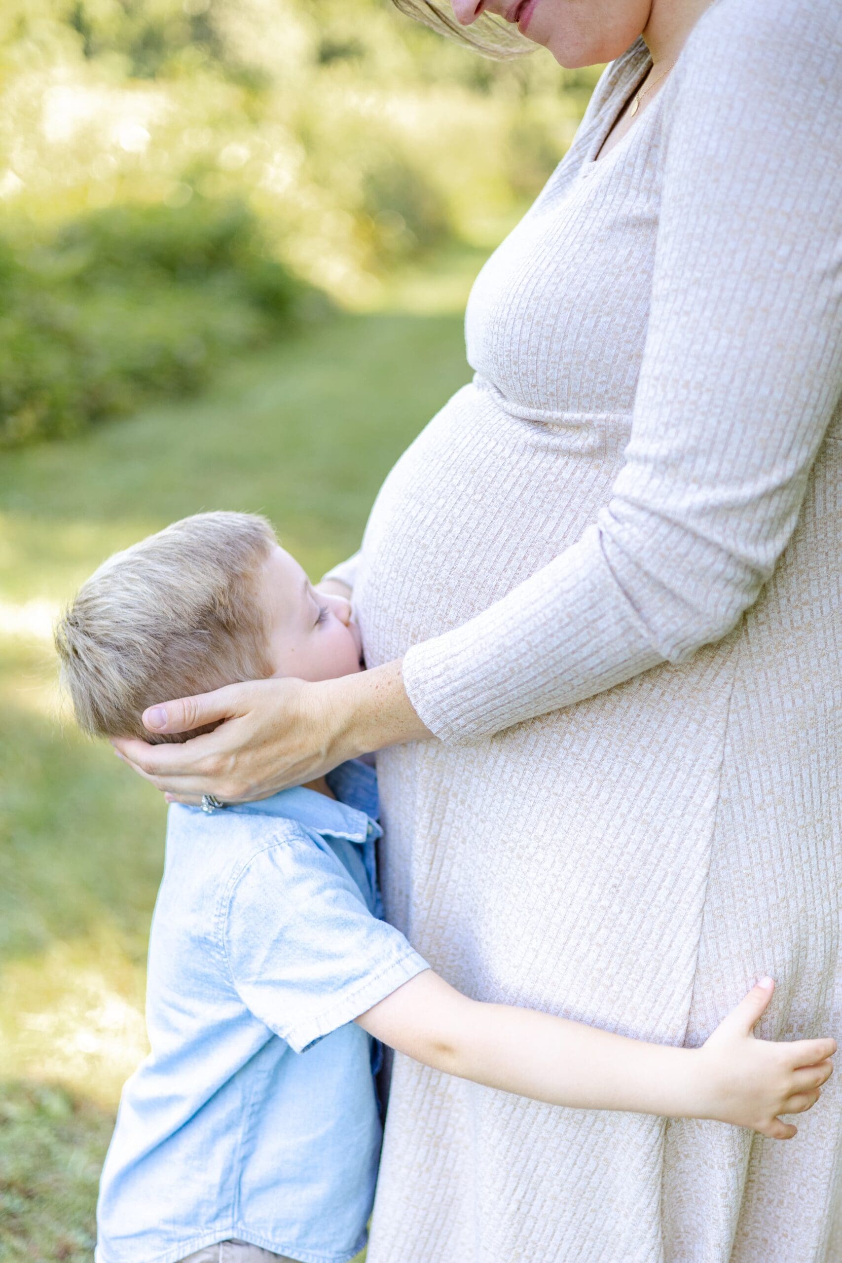 Young boy in blue kisses his mother's pregnant belly. She is wearing a tan dress and they are in a grassy field. She has her hands placed on his head.