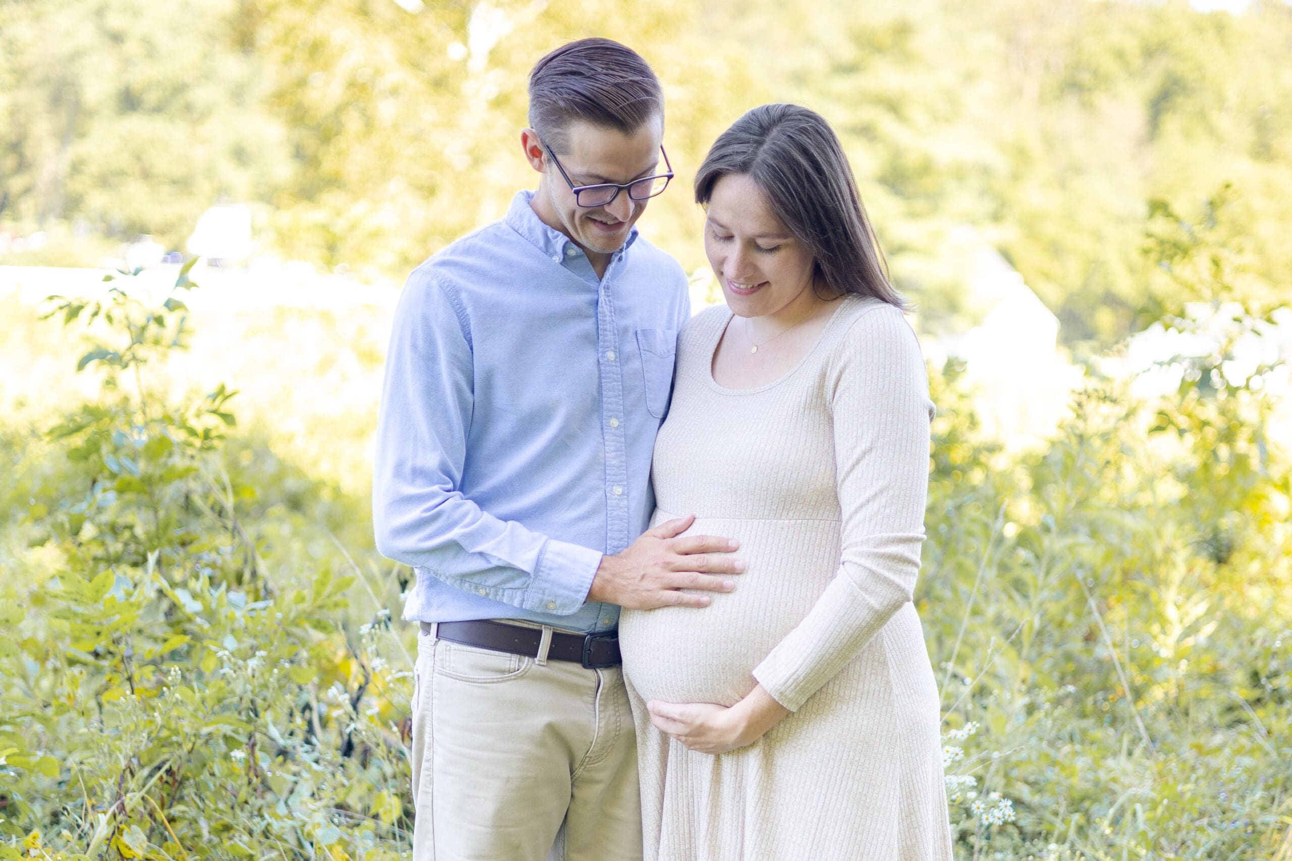 A couple stands close, the man gently placing his hand on the woman's pregnant belly. They are holding hands, conveying a sense of love and anticipation.