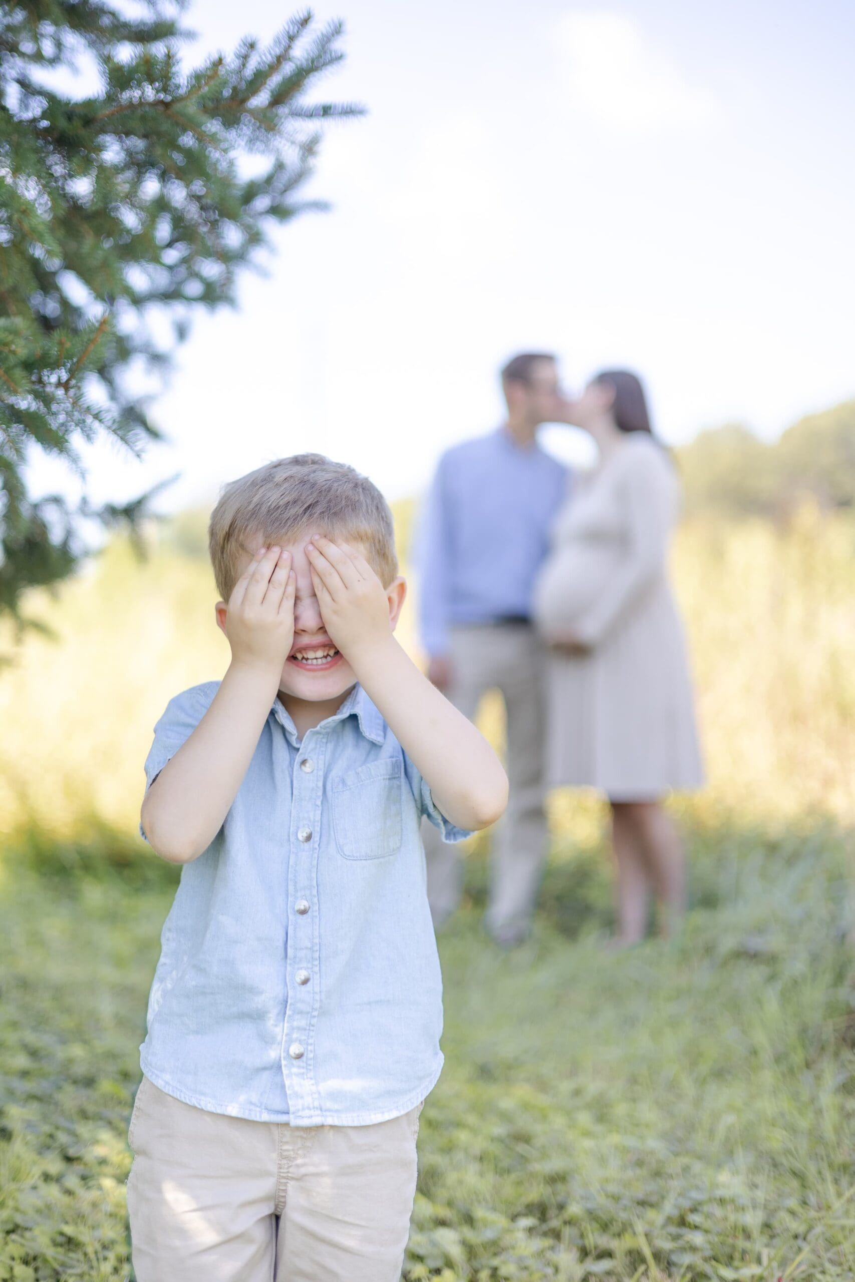 Little boy in blue stands focused in the foreground while he covers his hands to his eyes and smiles. In the background, a man and woman kiss, holding the woman's pregnant belly.