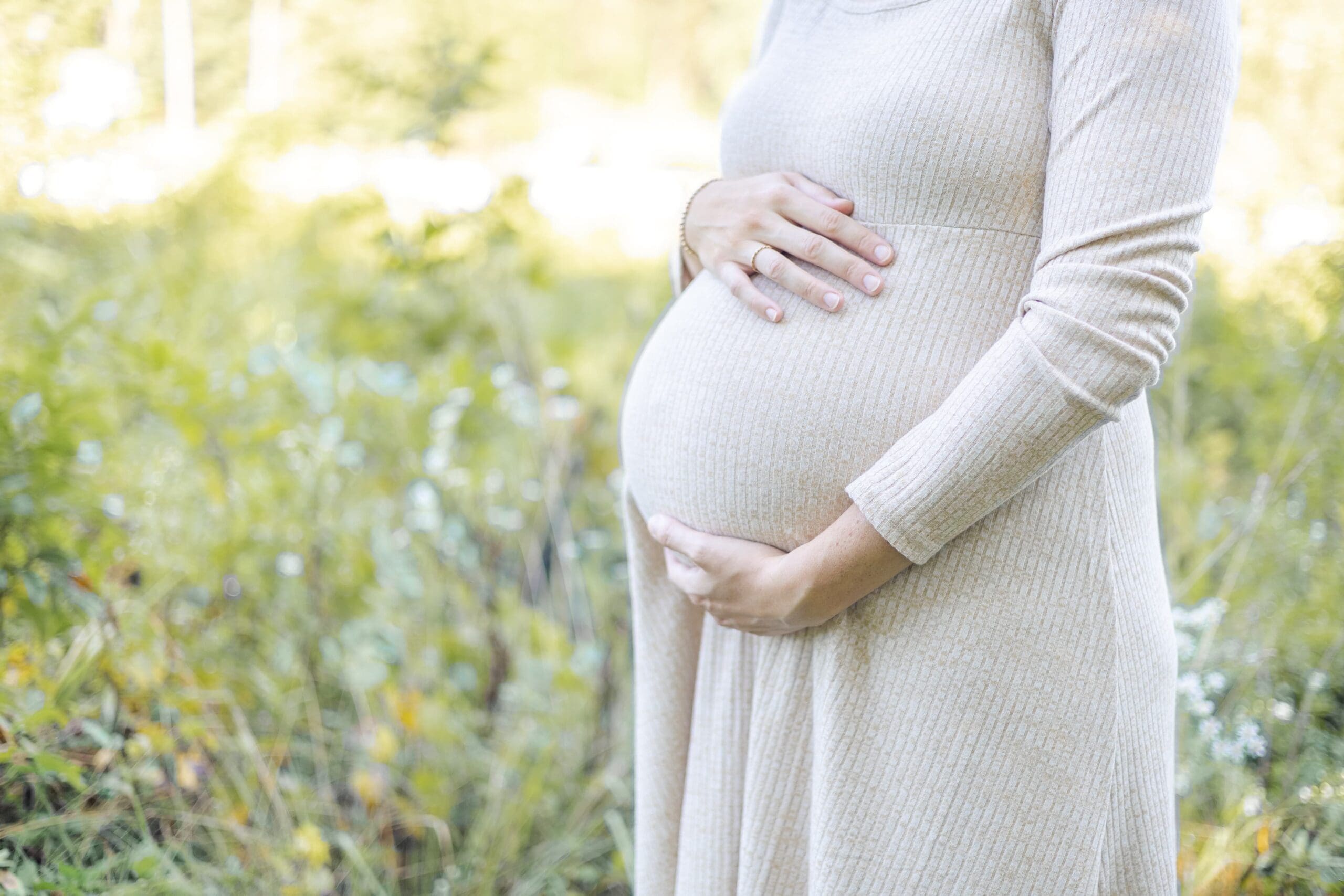 woman in tan dress places her hands on her pregnant belly. She conveys a sense of love and anticipation.