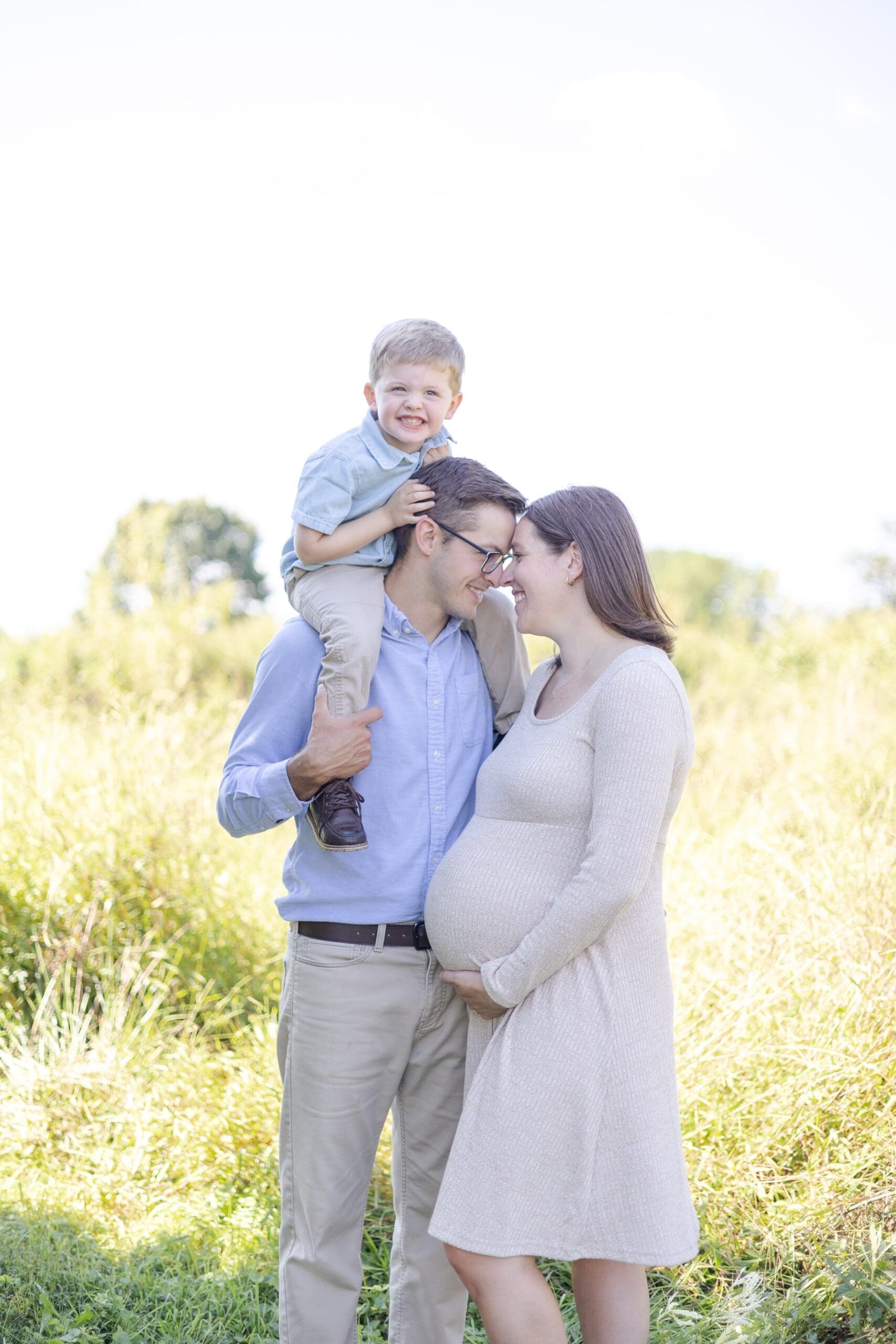 A pregnant woman and a man stand in a sunlit field, smiling. The man carries a young child on his shoulders, creating a warm family moment.
