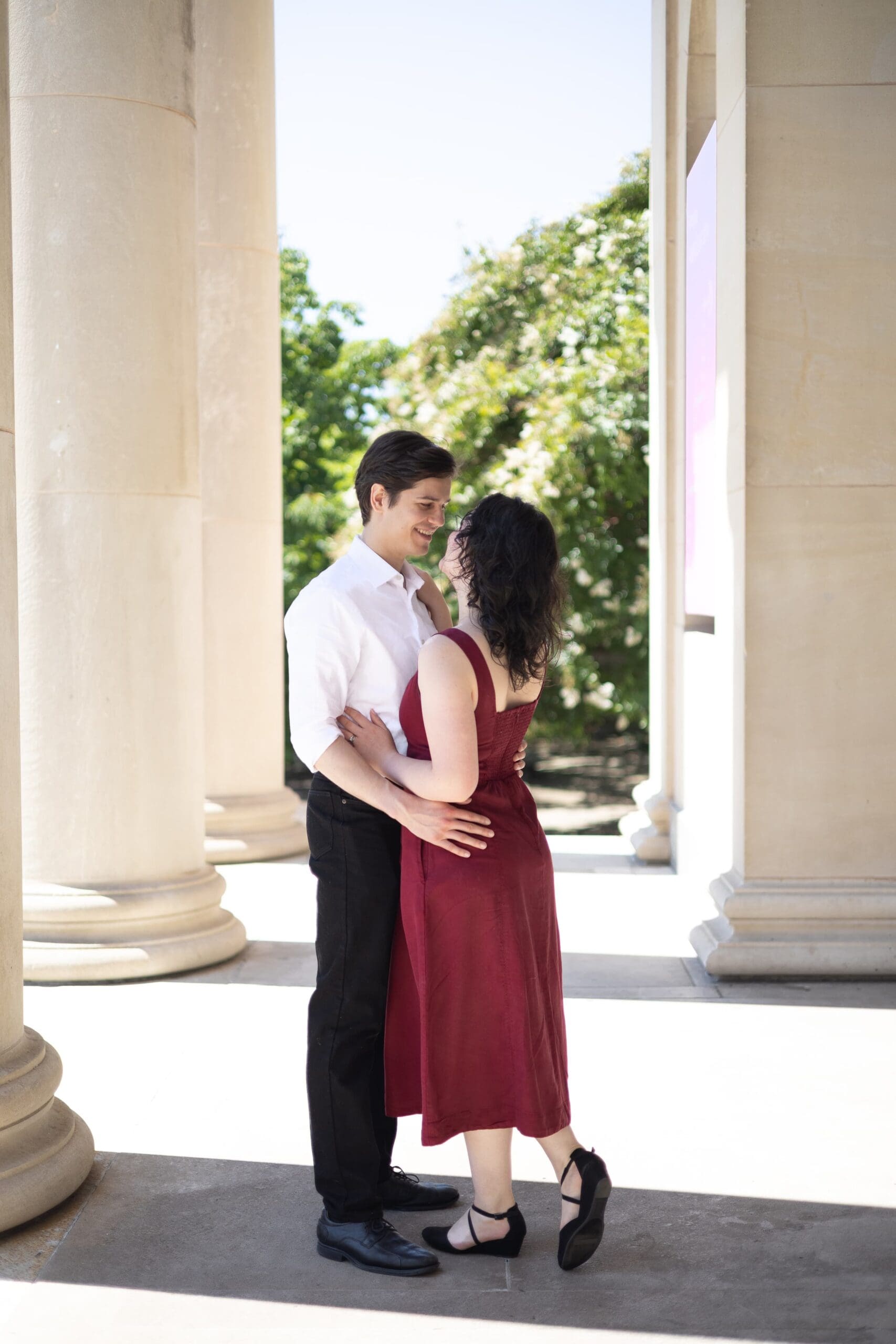 Couple hold each other close outside the Baltimore Museum of Art. Stone pillars surround them as the sun lights up the space. Photo taken by Lauren Beth Photos.