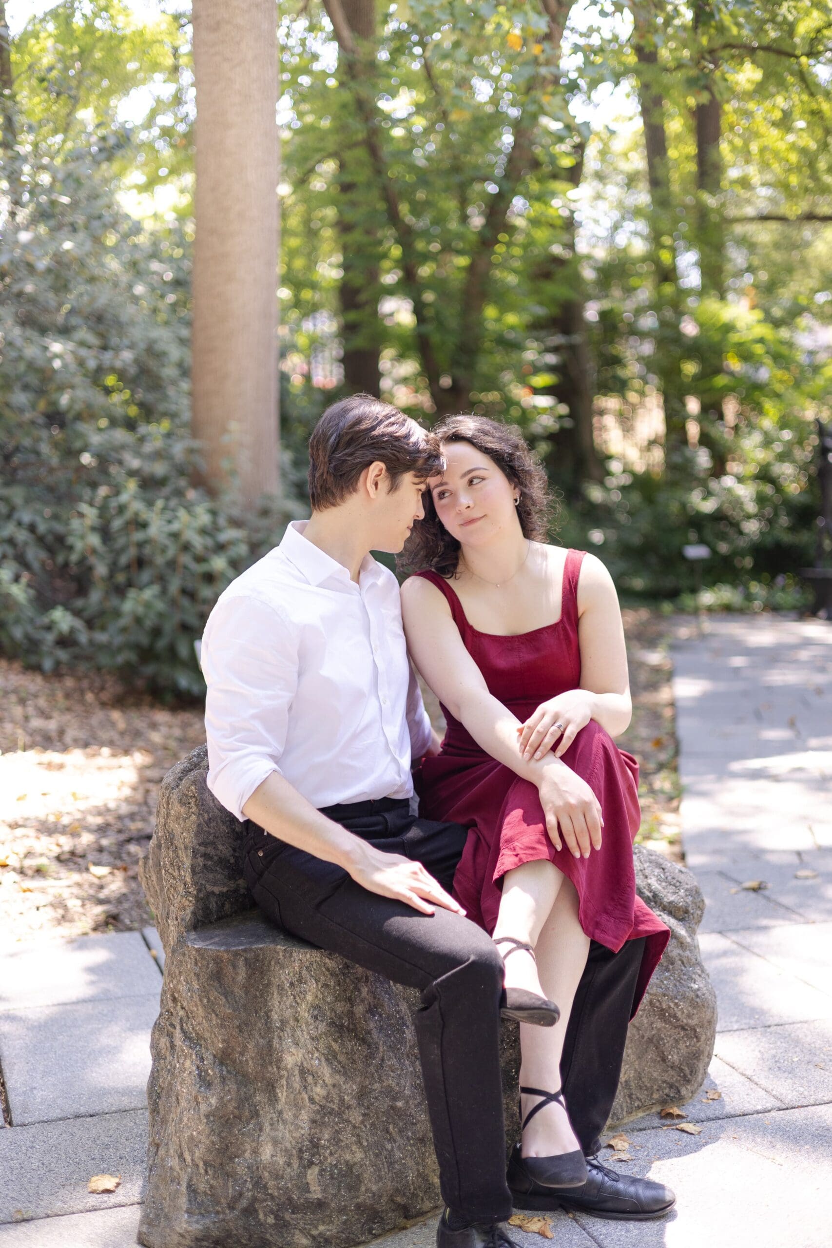 A couple sits closely on a rock in The Baltimore Museum of Art garden, surrounded by trees. They gaze affectionately at each other, creating a serene, romantic atmosphere.